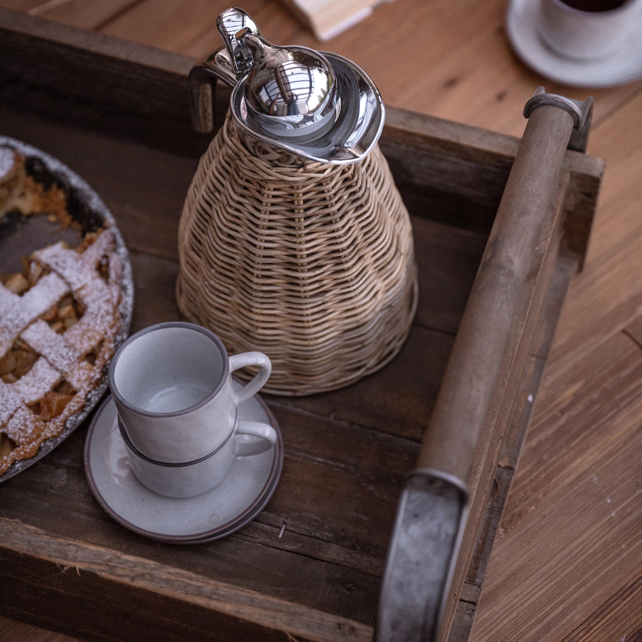 Holztablett mit geflochtenem Krug und zwei weissen Tassen neben Kuchen auf Holztisch