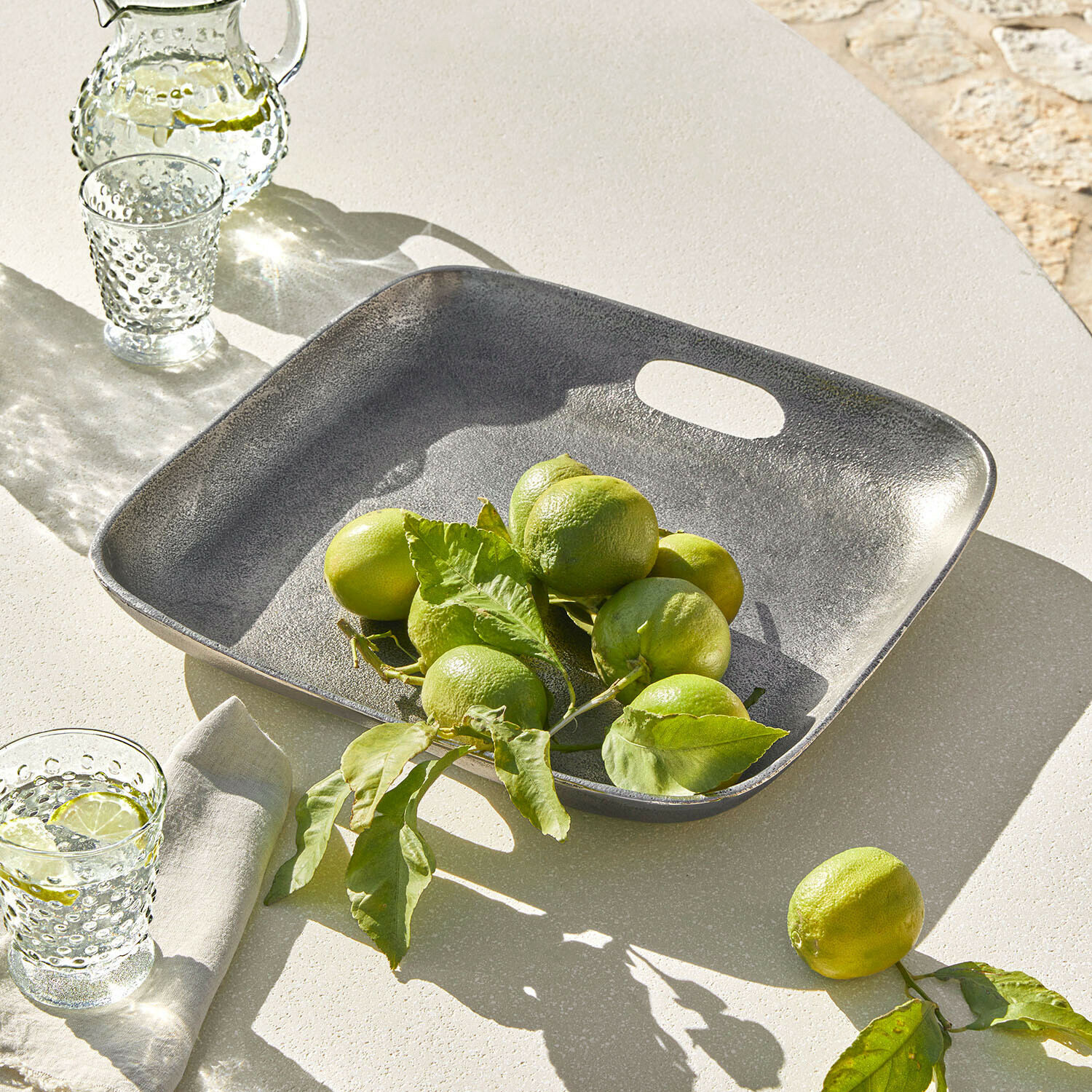 Quadratische graue Metallplatte mit grünen Limetten auf hellem Tisch, sommerliche Tischdekoration