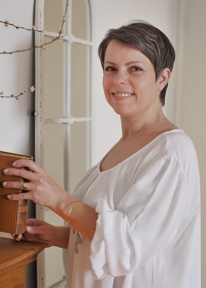 Frau in weisser Baumwollbluse hält altes Buch an einem Holzregal in hellem Raum.