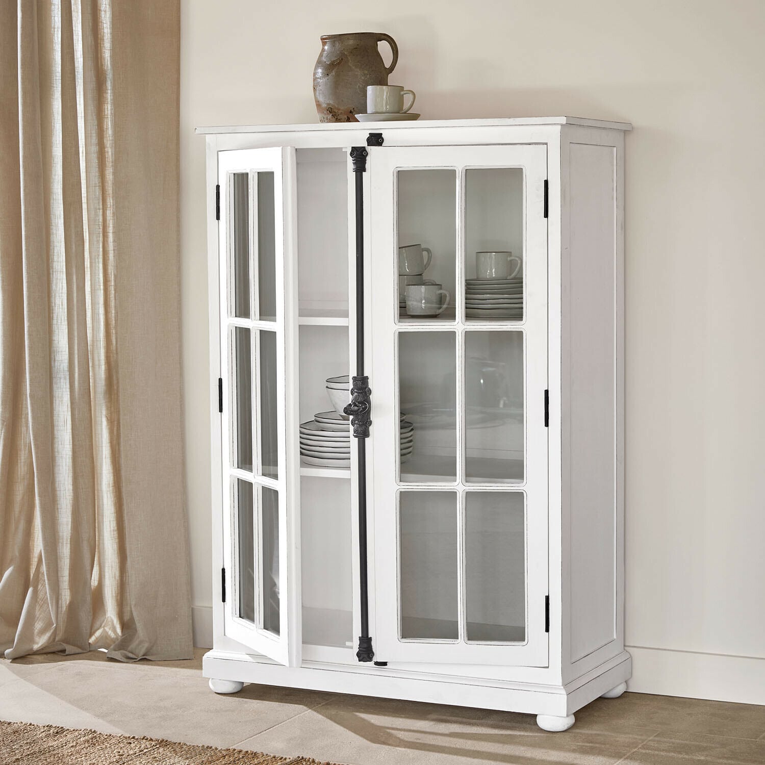 Schrank Salivonne Weisse Holzvitrine mit verglasten Türen und schwarzen Metallgriffen im Vintage-Stil in hellem Raum
