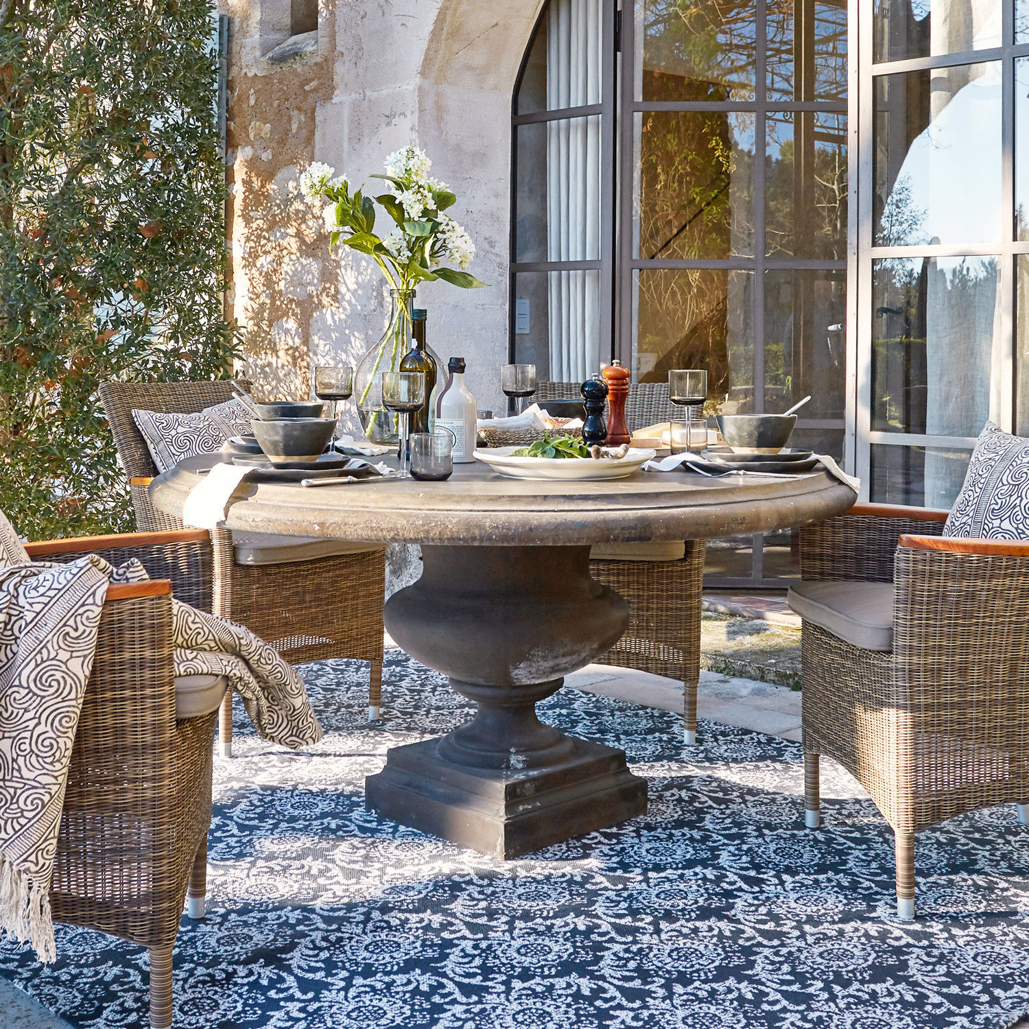 Tisch Monterey Runder Steintisch mit schwarzem Sockel und Rattanstühlen auf der Terrasse mit gemustertem Teppich