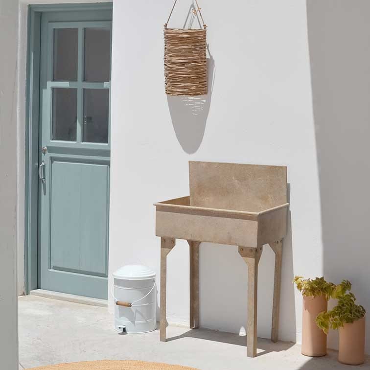 Beige Steintopf auf Beinen vor weisser Wand mit blauer Tür und hängendem Weidenkorb