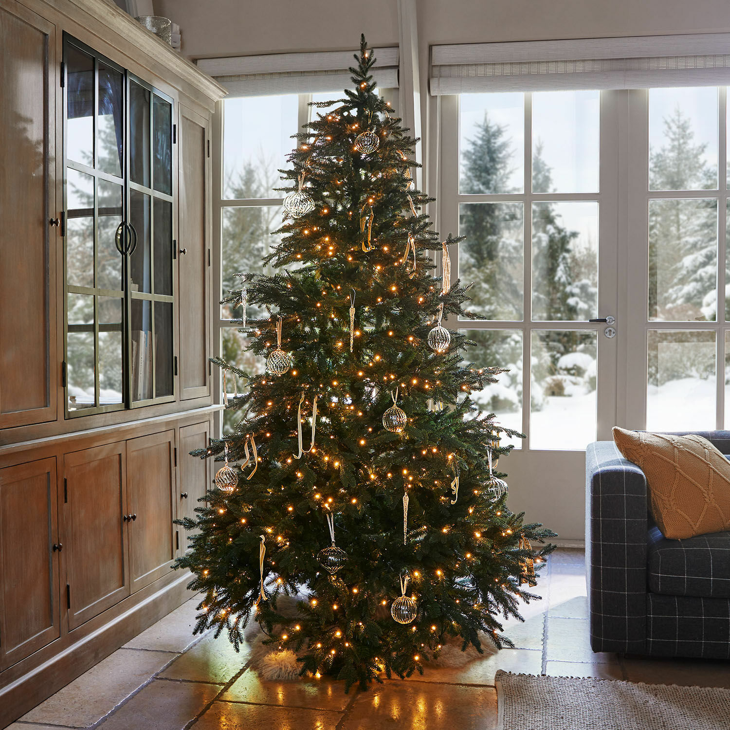 Weihnachtsbaum Celindor Beleuchteter Weihnachtsbaum mit grünen Zweigen und gläsernen Kugeln in einem gemütlichen Wohnzimmer mit Schneeblick