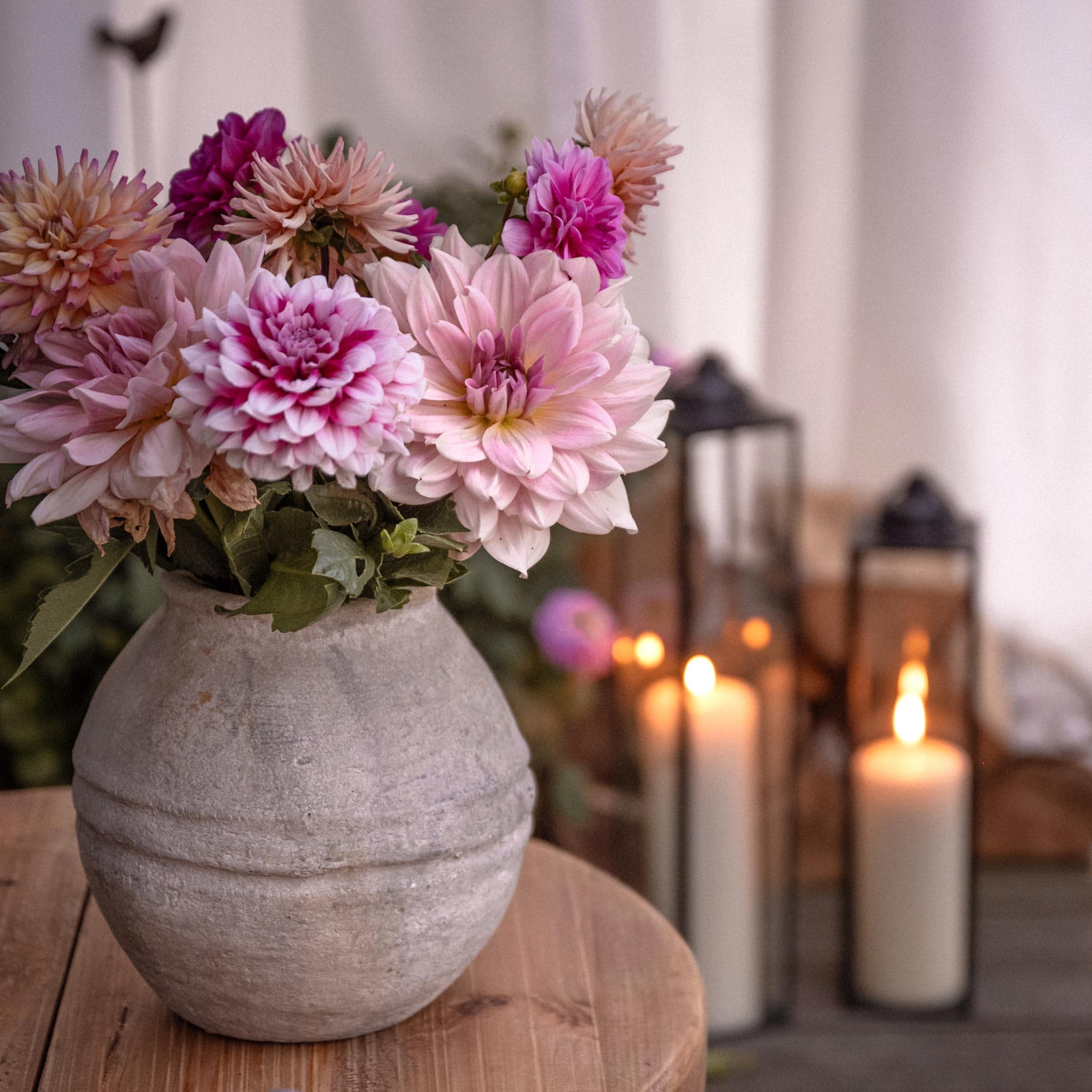 Graue Dekovase mit rosa-violetten Blumen auf Holztisch, brennende Laternen im Hintergrund