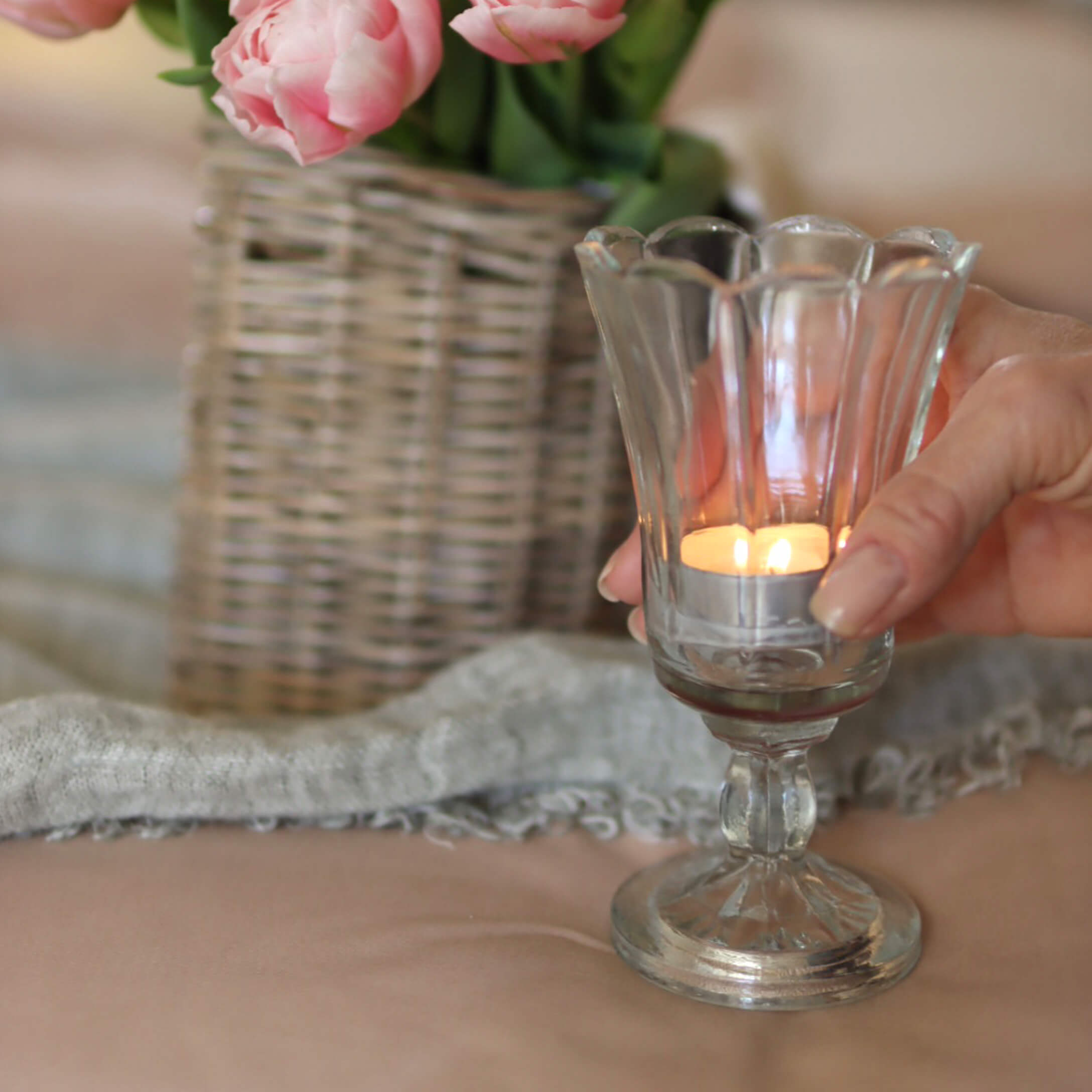 Glas-Teelichthalter auf Fuss mit brennender Kerze, rosa Blumen in Korbvase im Hintergrund
