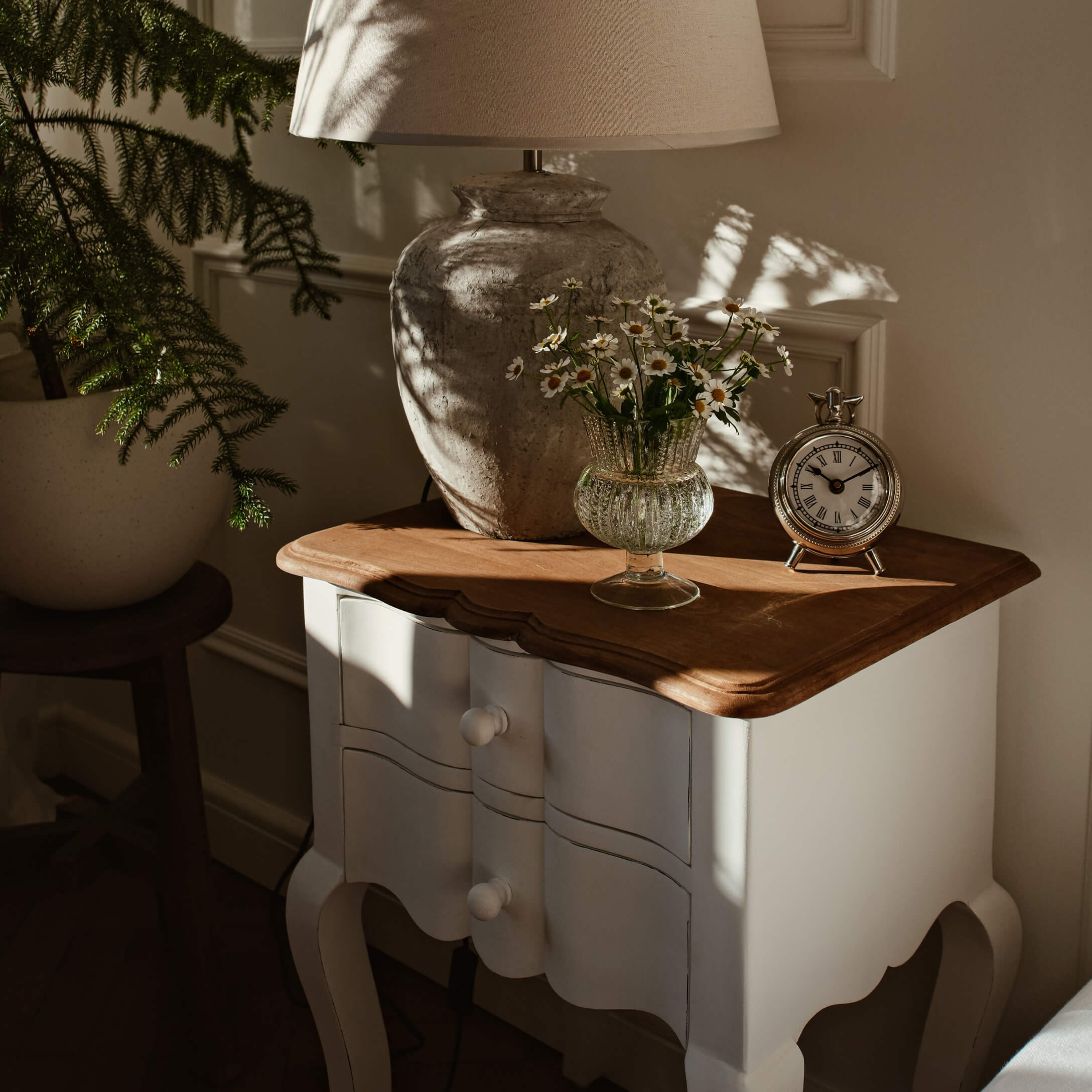 Weisse Holzkommode mit brauner Platte, Lampe, Glasvase mit Blumen und silberner Uhr im Sonnenlicht