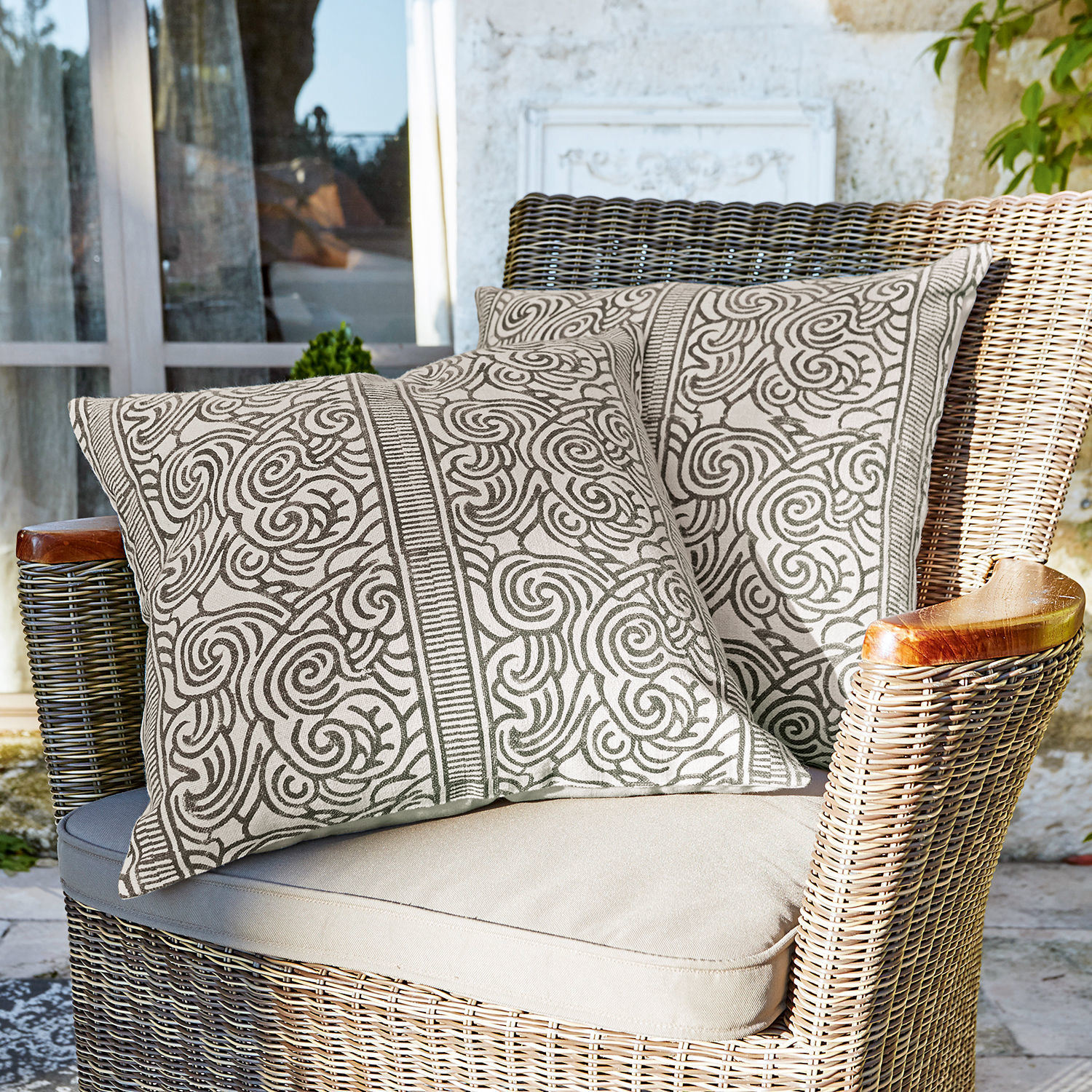 Kissenhülle 2er Set Ensis Zwei dekorative Baumwollkissen in Beige mit braunem Muster auf einem Rattansessel auf der Terrasse