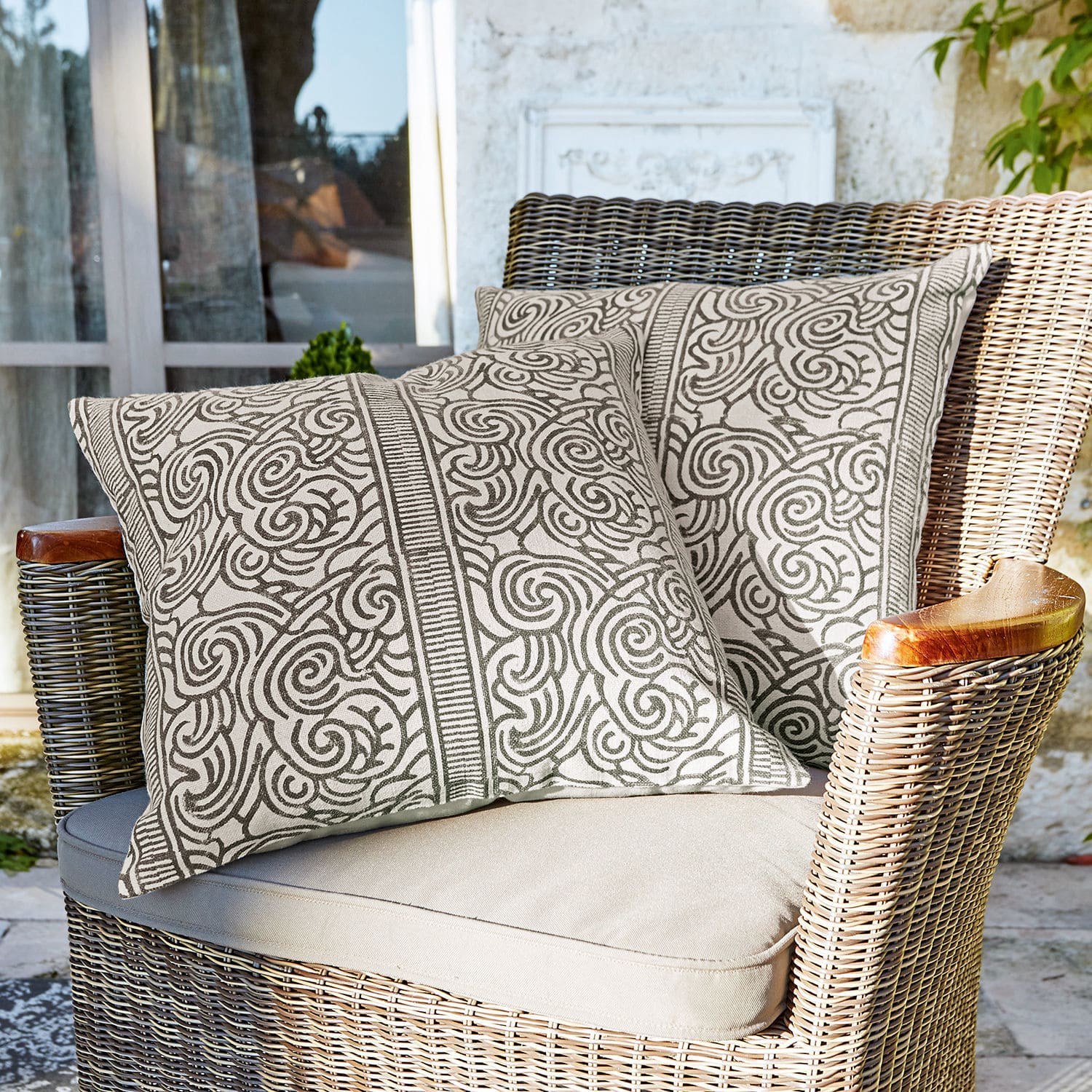 Kissenhülle 2er Set Ensis Zwei dekorative Baumwollkissen in Beige mit braunem Muster auf einem Rattansessel auf der Terrasse
