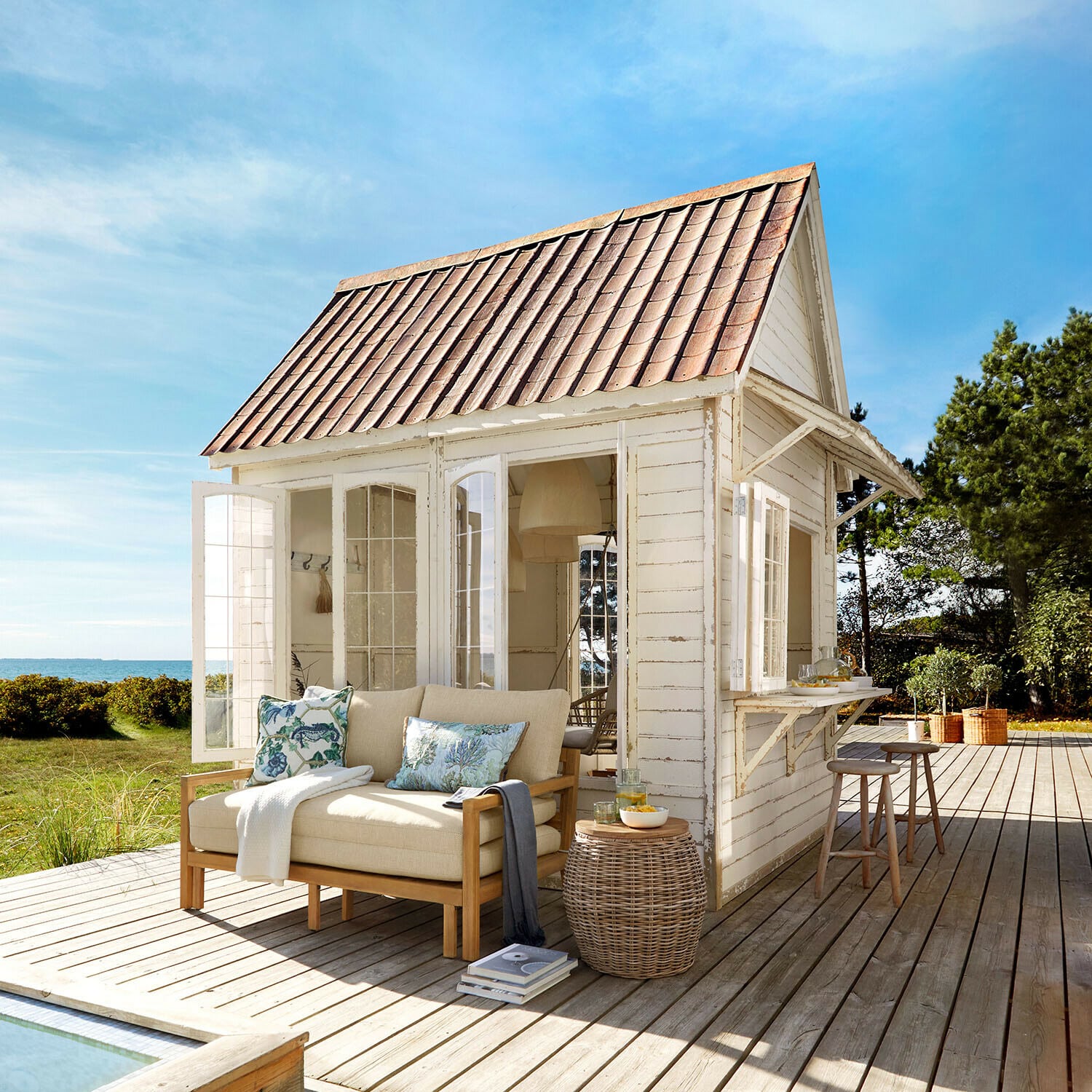 Gartenhaus Stormhavn Weisses Holzhaus mit rotem Dach und beigem Sofa auf Holzterrasse mit Meerblick