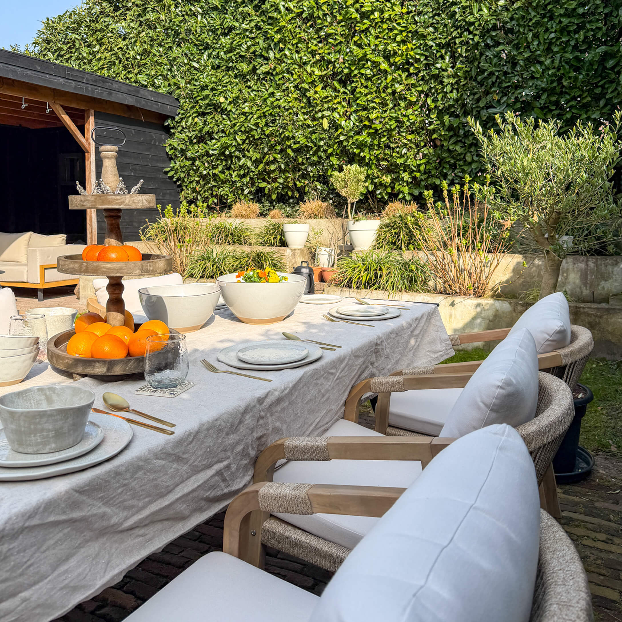 Gartentisch mit weissem Leinentuch, Keramikgeschirr und hölzernem Obstständer bei Sonnenschein