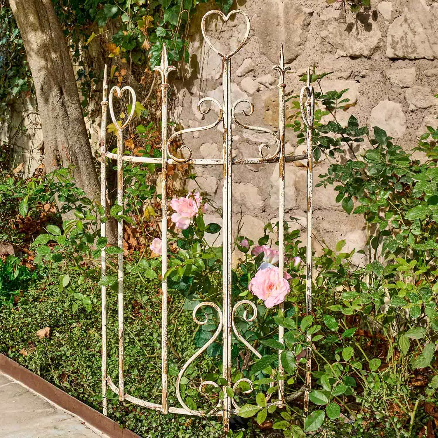 Weisses Metall-Gartengitter mit Herzmotiven und rosa Rosen vor Steinmauer im Garten