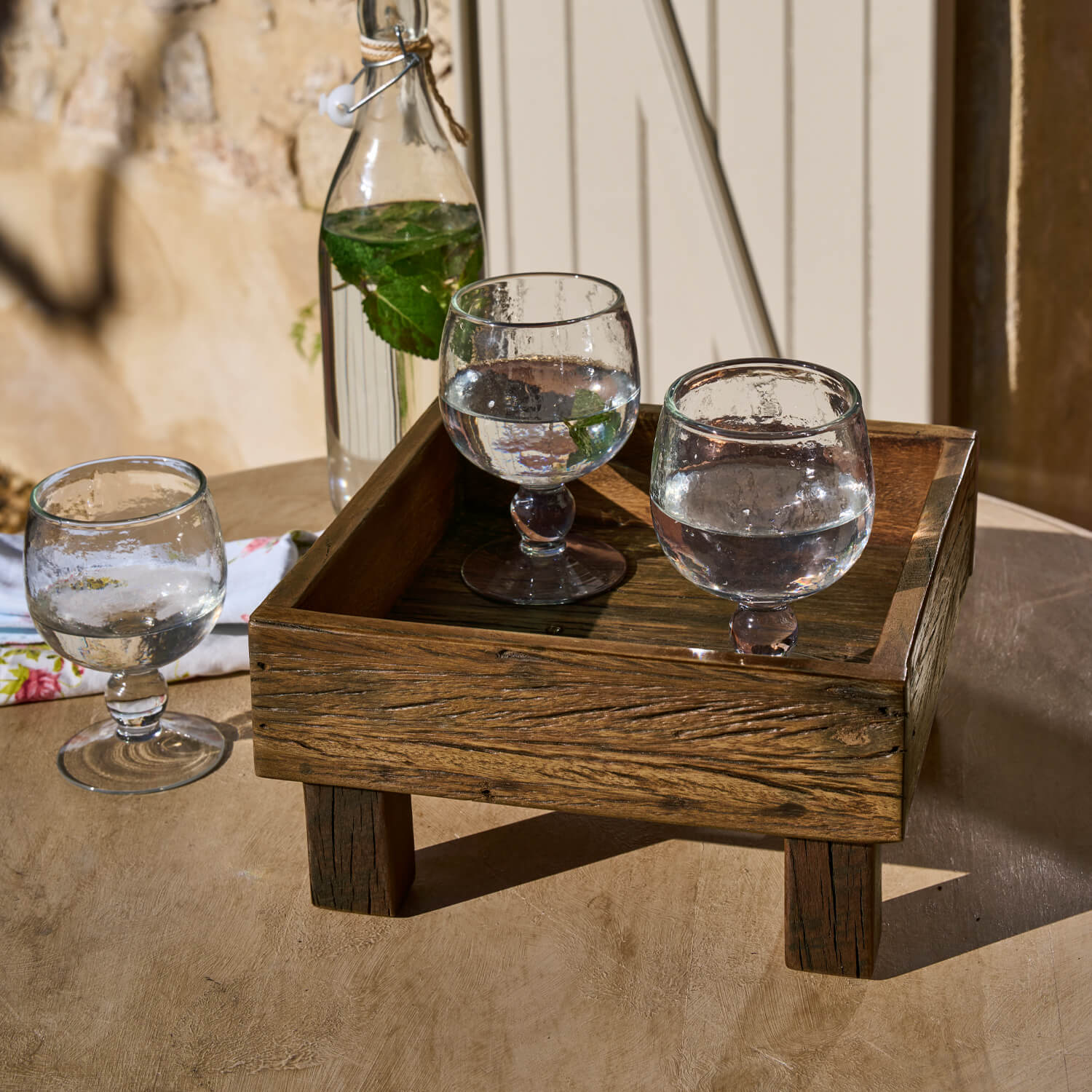 Holztablett auf Beinen mit drei Wassergläsern und Flasche mit Minzblättern auf Tisch
