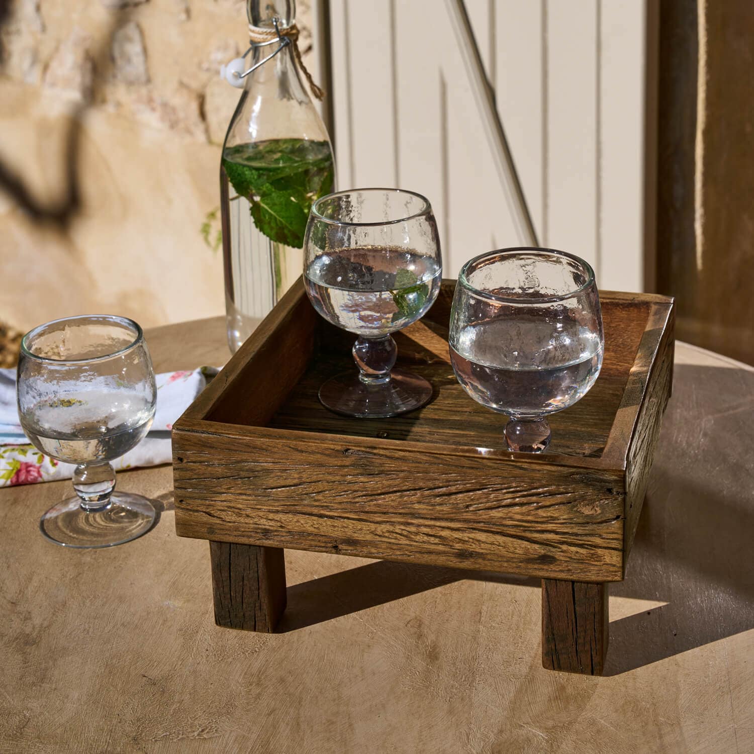 Holztablett auf Beinen mit drei Wassergläsern und Flasche mit Minzblättern auf Tisch