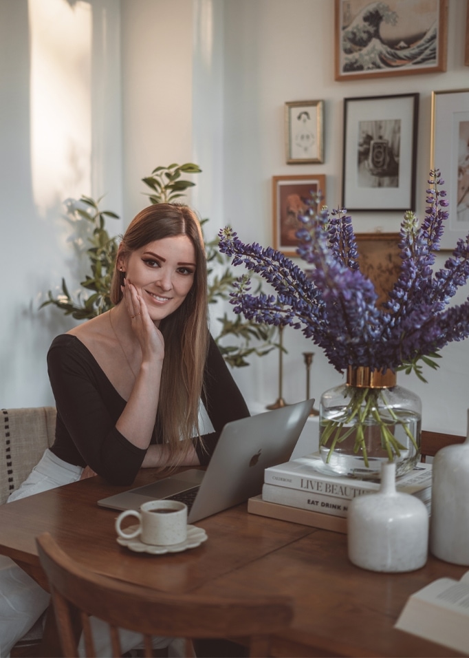Frau am Holztisch mit Laptop und Glasvase mit violetten Blumen in hellem Raum