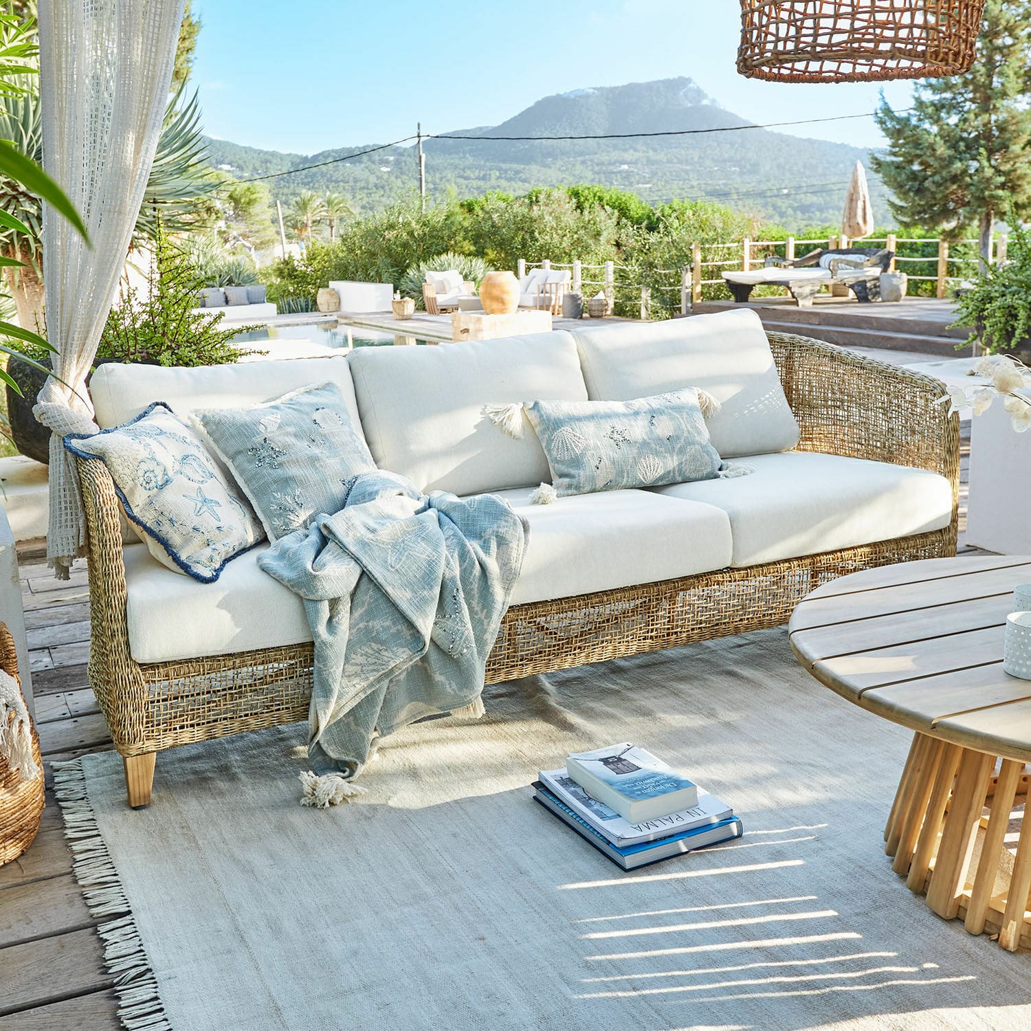 Sofa Clarivaux Rattansofa mit cremefarbenen Kissen und blauem Plaid auf der Terrasse mit Bergsicht