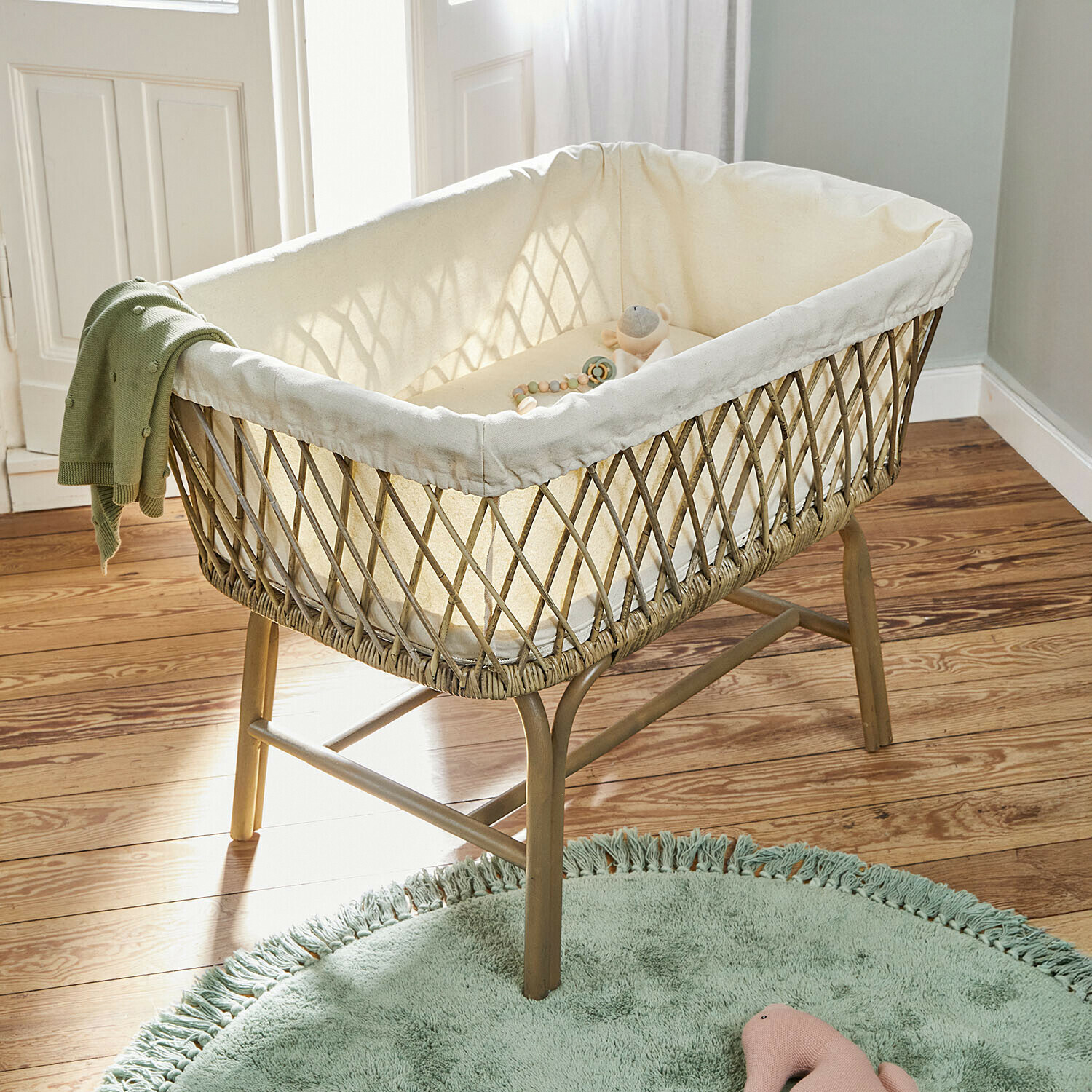 Babybettchen Nuvinas Kinderkorb aus Korbgeflecht mit cremefarbiger Polsterung und grünem Strickdecke auf Holzbeinen im Kinderzimmer