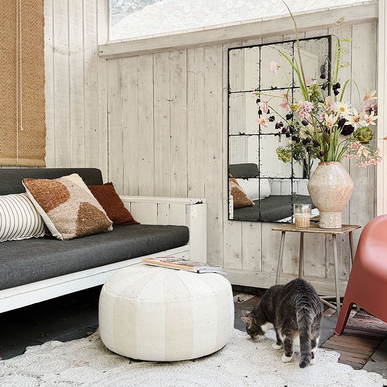 Gemütliches Wohnzimmer mit weisser Holzverkleidung, grauem Sofa, weissem Pouf und Blumenarrangement