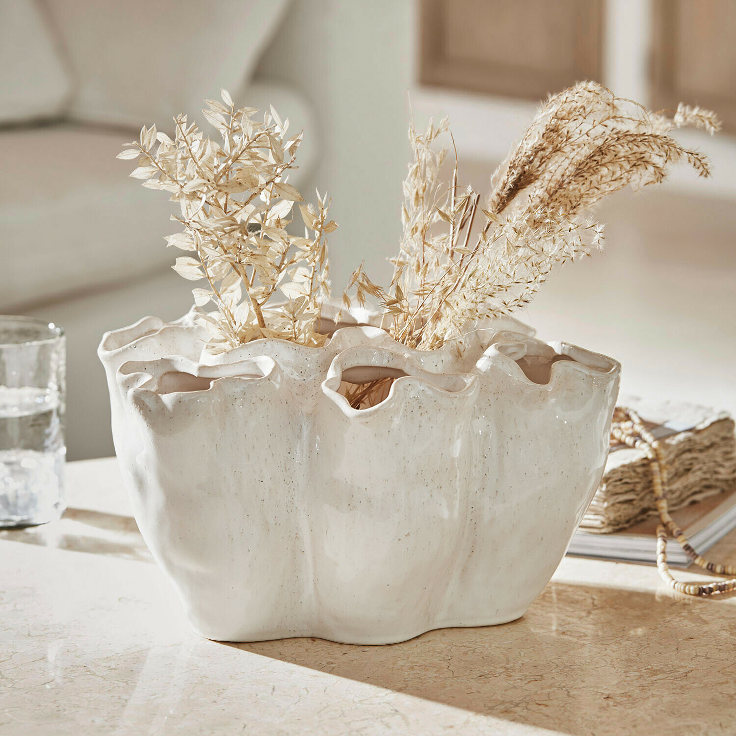Weisse Keramikvase mit welligem Rand und getrockneten Pflanzen auf dekorativem Tisch