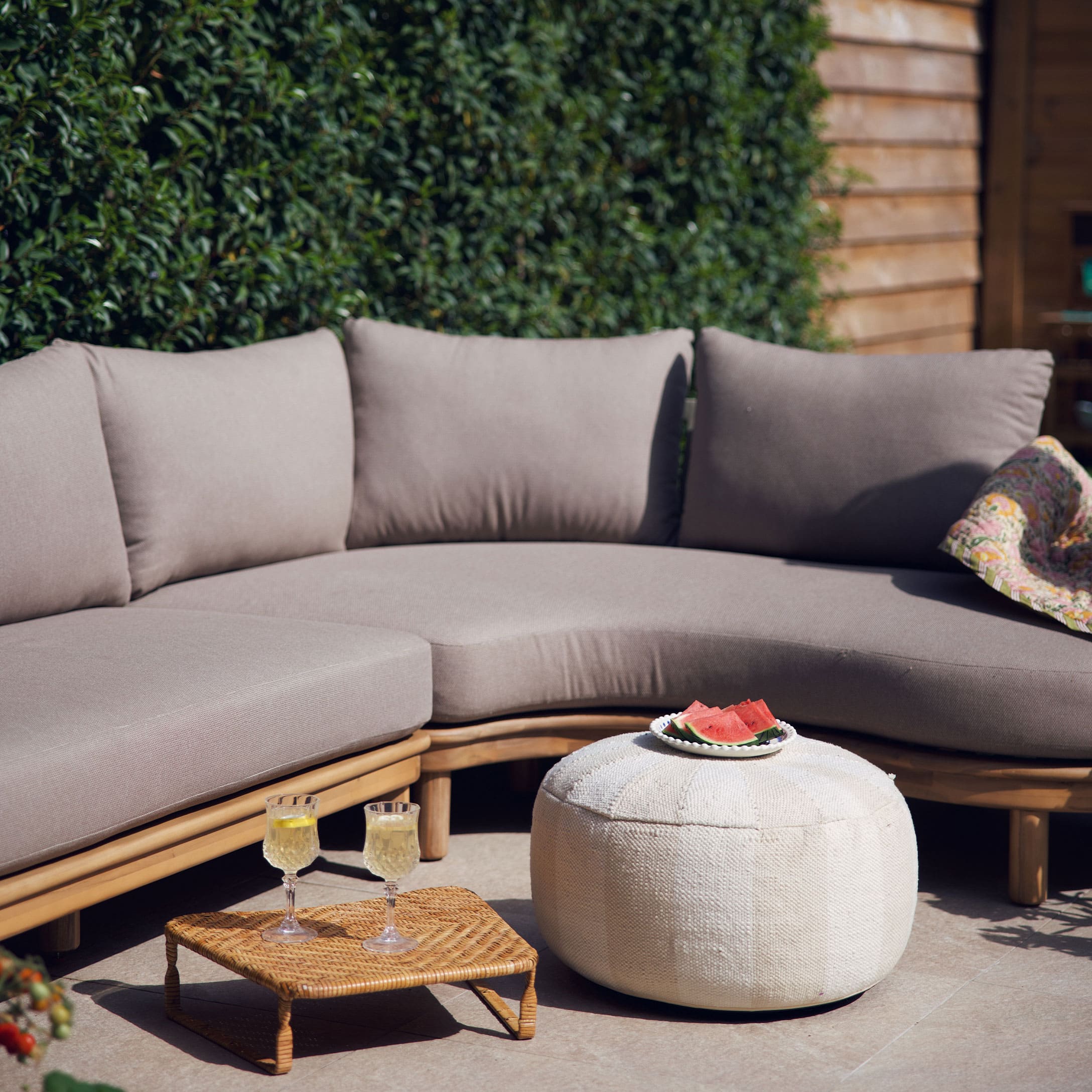 Beige Sofa mit Kissen auf Holzrahmen und weissem Pouf auf Terrasse mit sommerlicher Dekoration