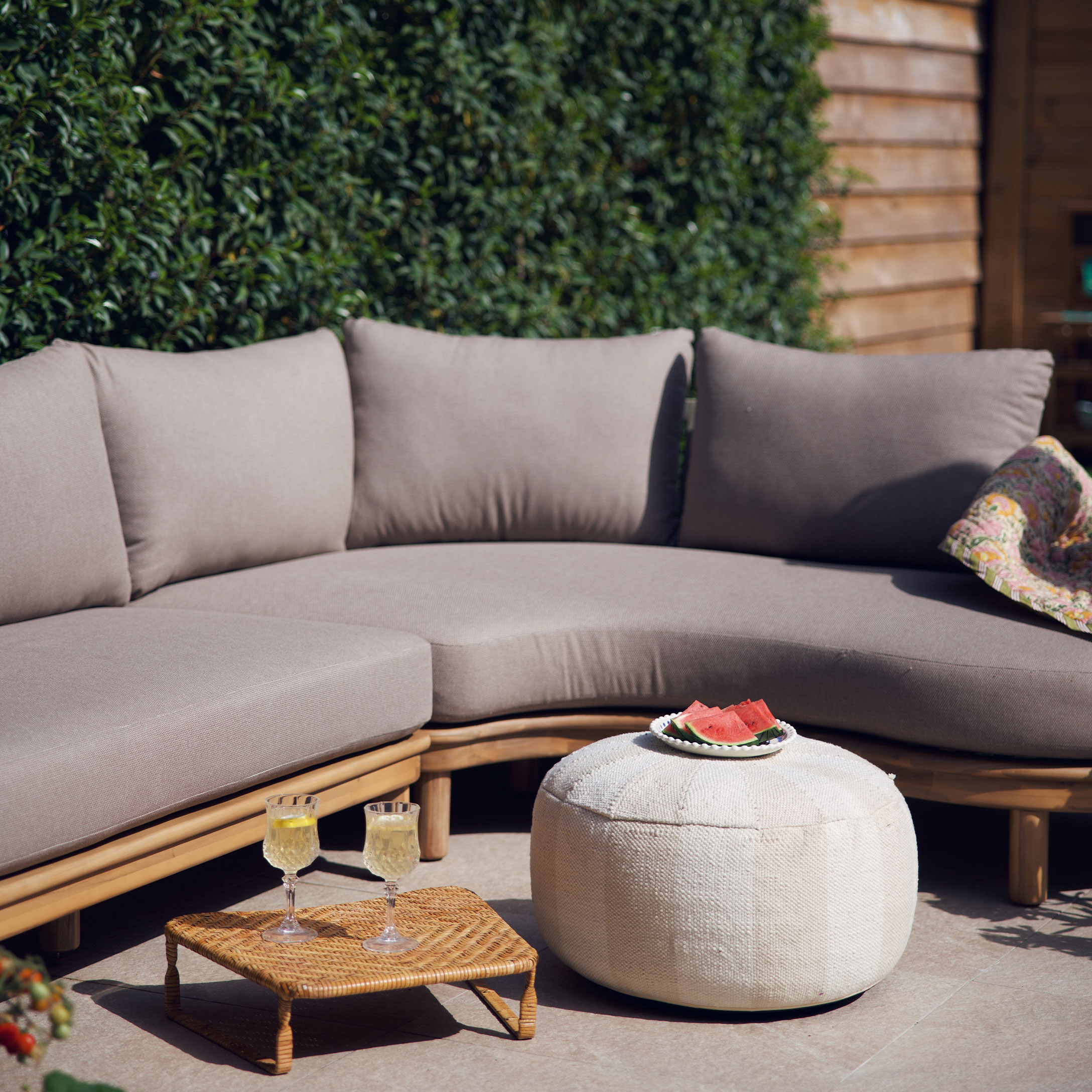 Beige Sofa mit Kissen auf Holzrahmen und weissem Pouf auf Terrasse mit sommerlicher Dekoration