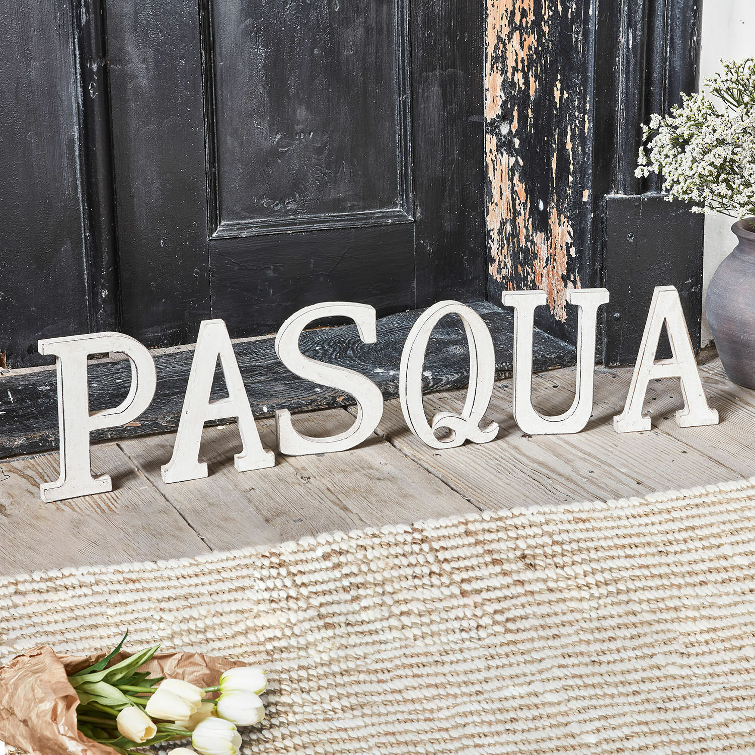 Deko-Buchstaben PASQUA Weisse Holzbuchstaben als Dekoration mit dem Schriftzug Pasqua auf Holzboden vor schwarzer Tür
