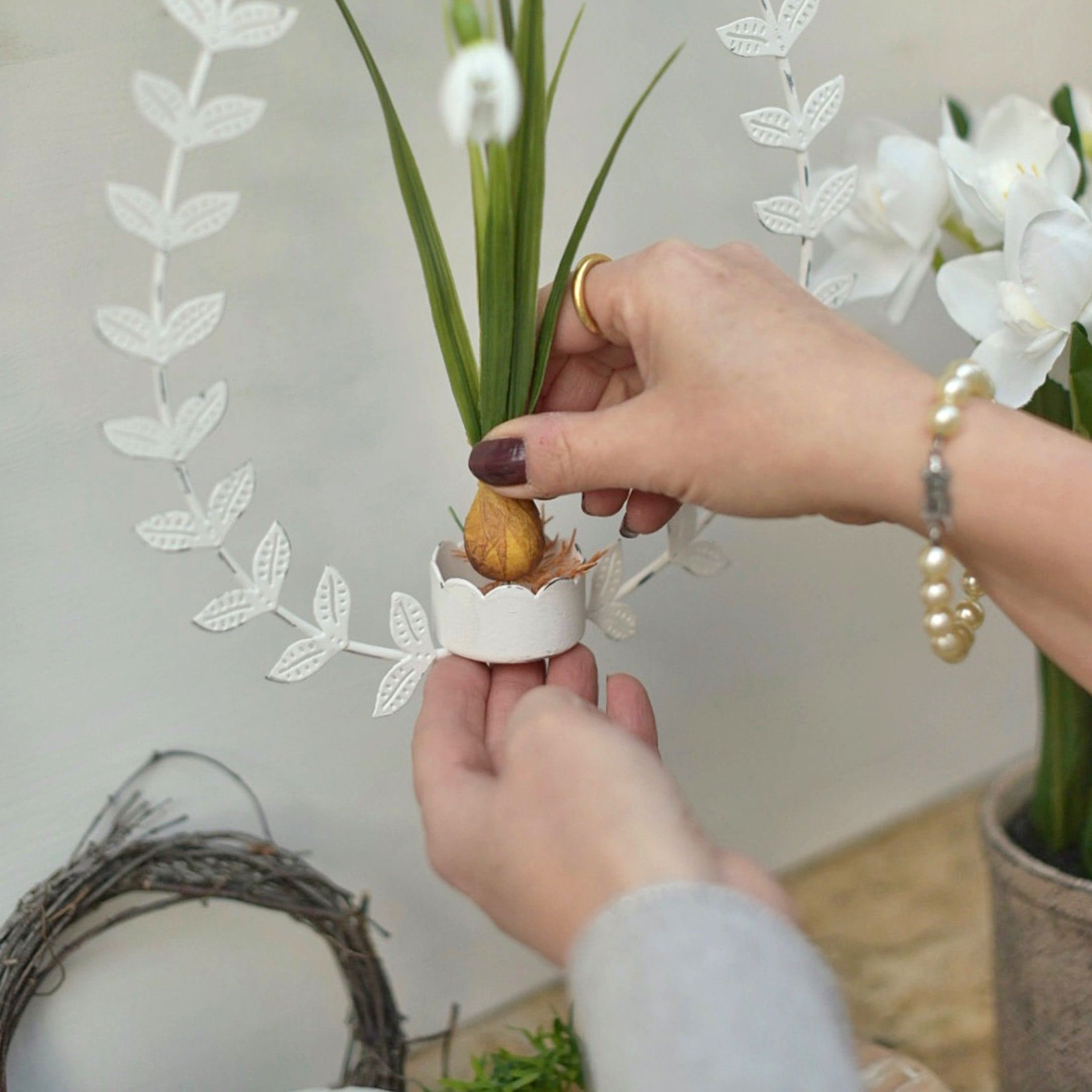 Hände setzen eine künstliche Blume in eine weisse Metall-Osterdekoration mit Blättern ein.