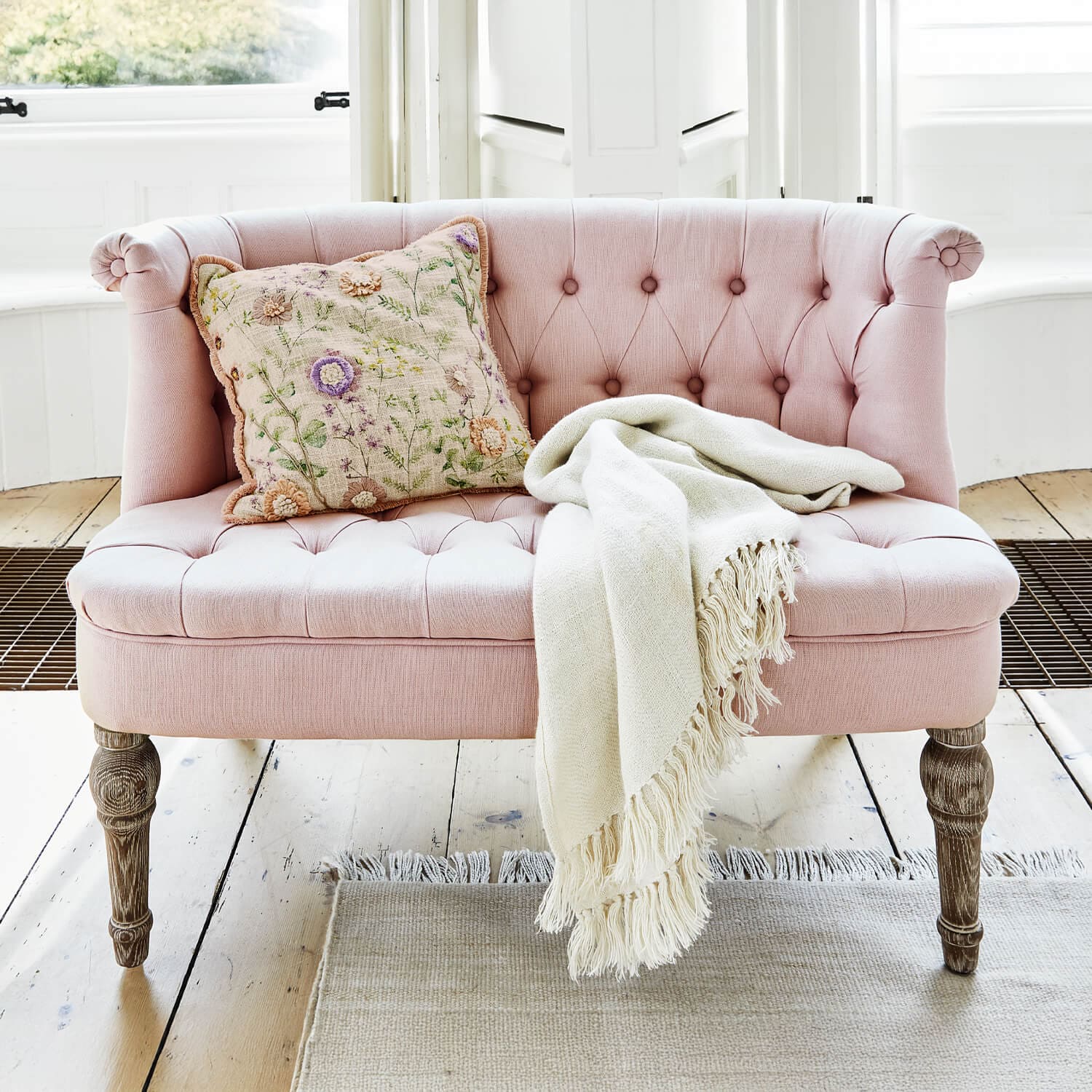 Rosa gepolstertes Sofa mit Holzbeinen, dekorativem Kissen und cremefarbenem Plaid auf hellem Boden