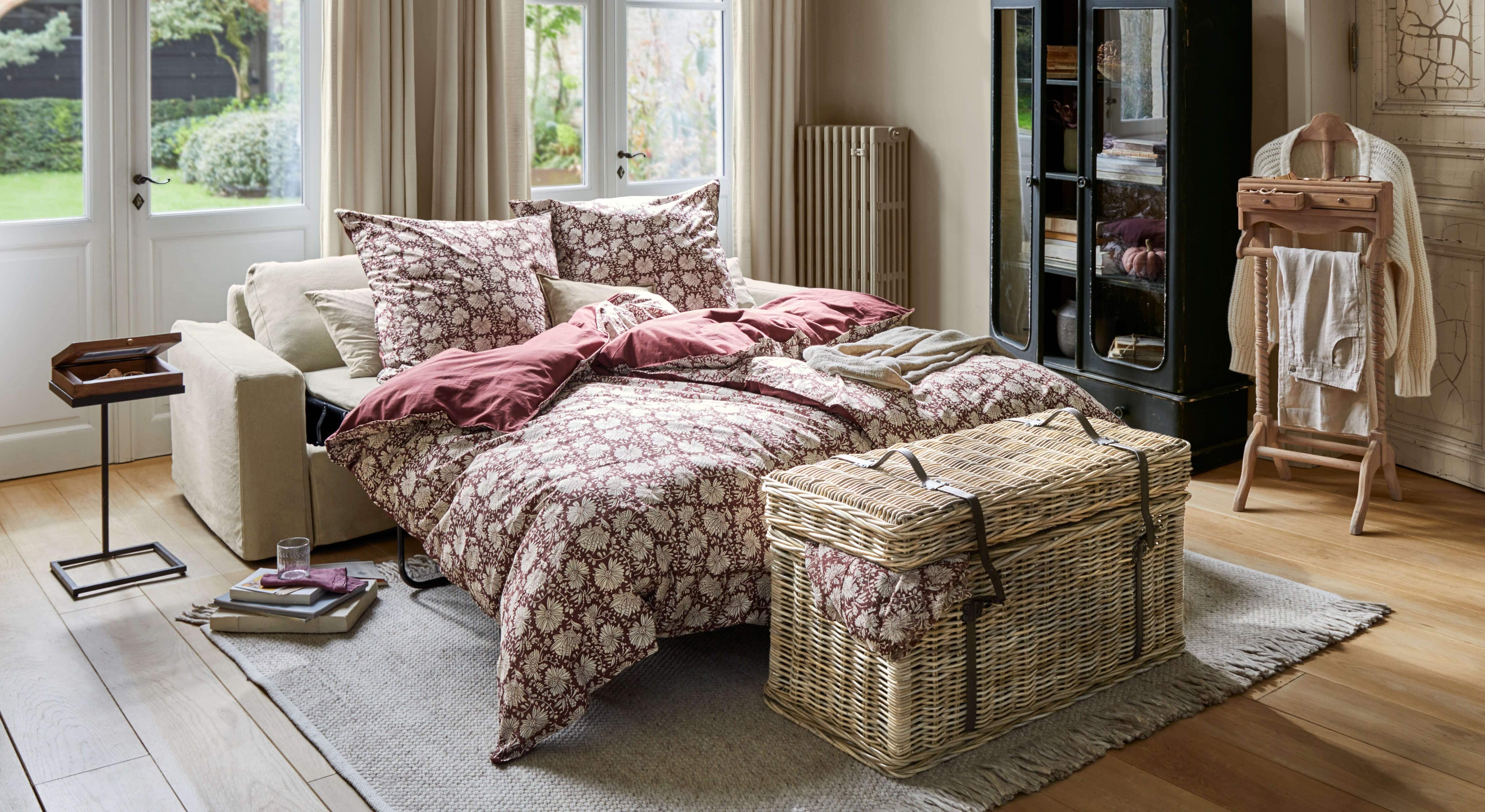 Ausziehbares Sofa mit beiger Polsterung und bordeauxfarbenem Blumenbettzeug in hellem Wohnzimmer