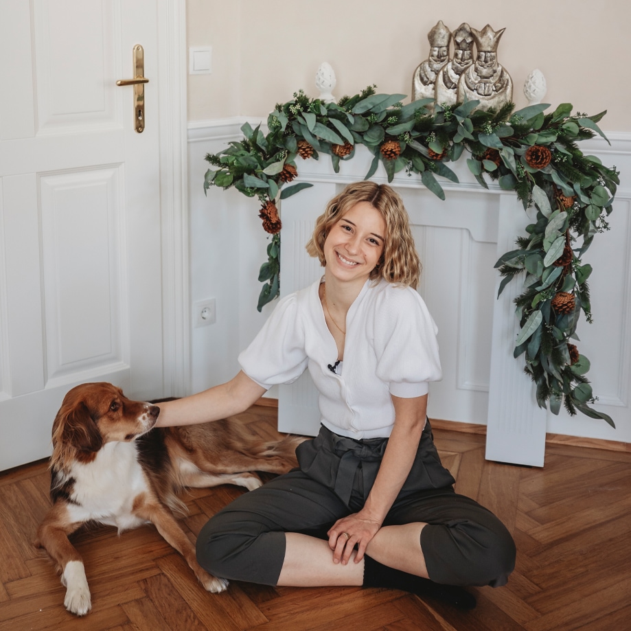 Junge Frau im weissen Pullover und dunkler Hose sitzt auf dem Holzboden mit Hund vor weihnachtlicher Dekoration aus grünen Blättern und Kiefernzapfen.