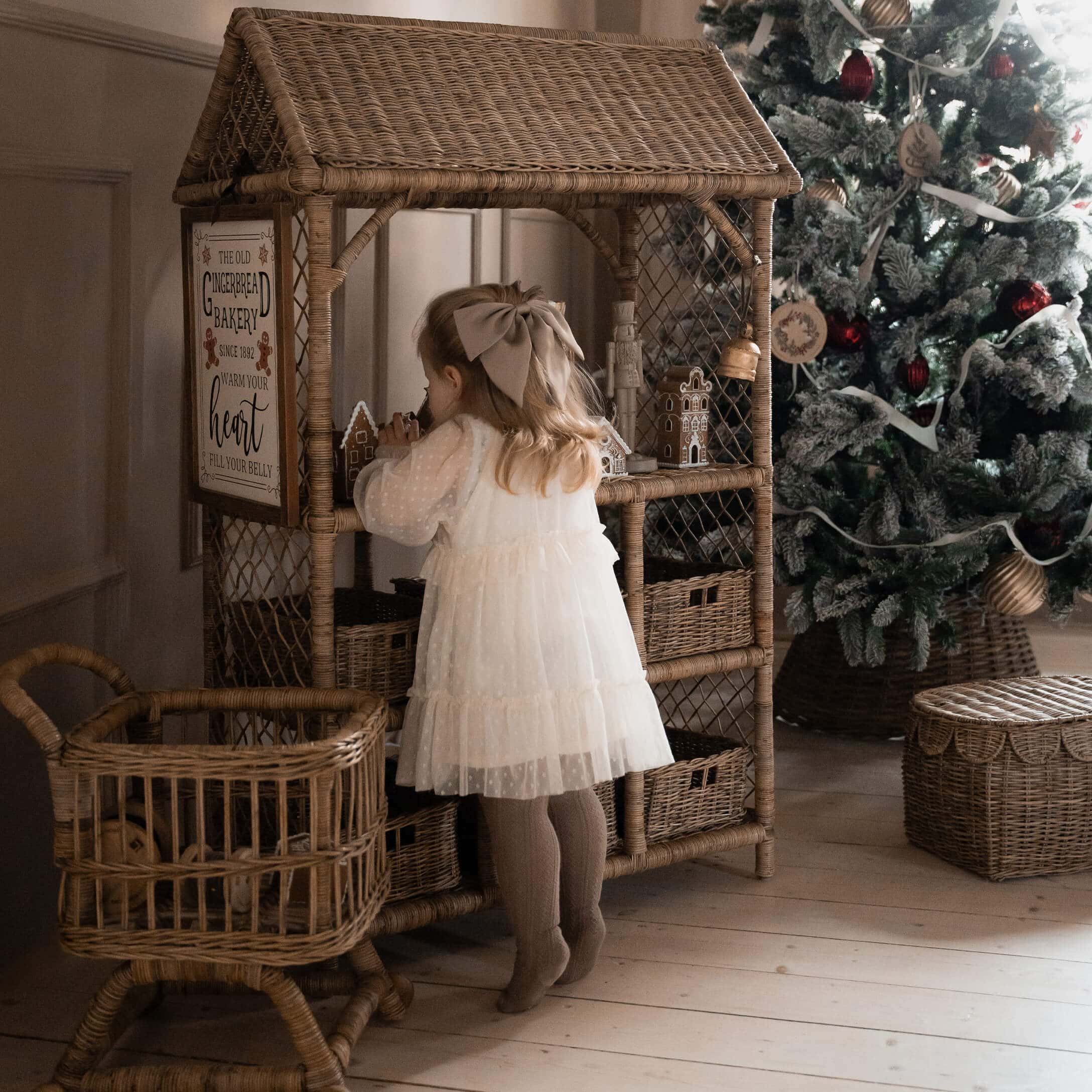 Kind in weissem Kleid vor einem Rattanschrank mit Lebkuchenhäusern neben geschmücktem Weihnachtsbaum