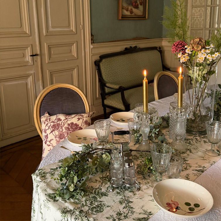 Eleganter Tisch mit weissem, grün gemustertem Tischtuch, brennenden Kerzen und Blumenarrangement im klassischen Raum