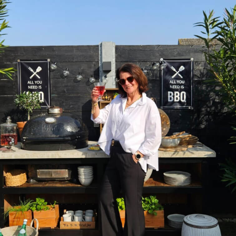 Frau in weisser Bluse und schwarzer Hose steht beim Aussen-Grill mit BBQ-Schild vor schwarzer Wand