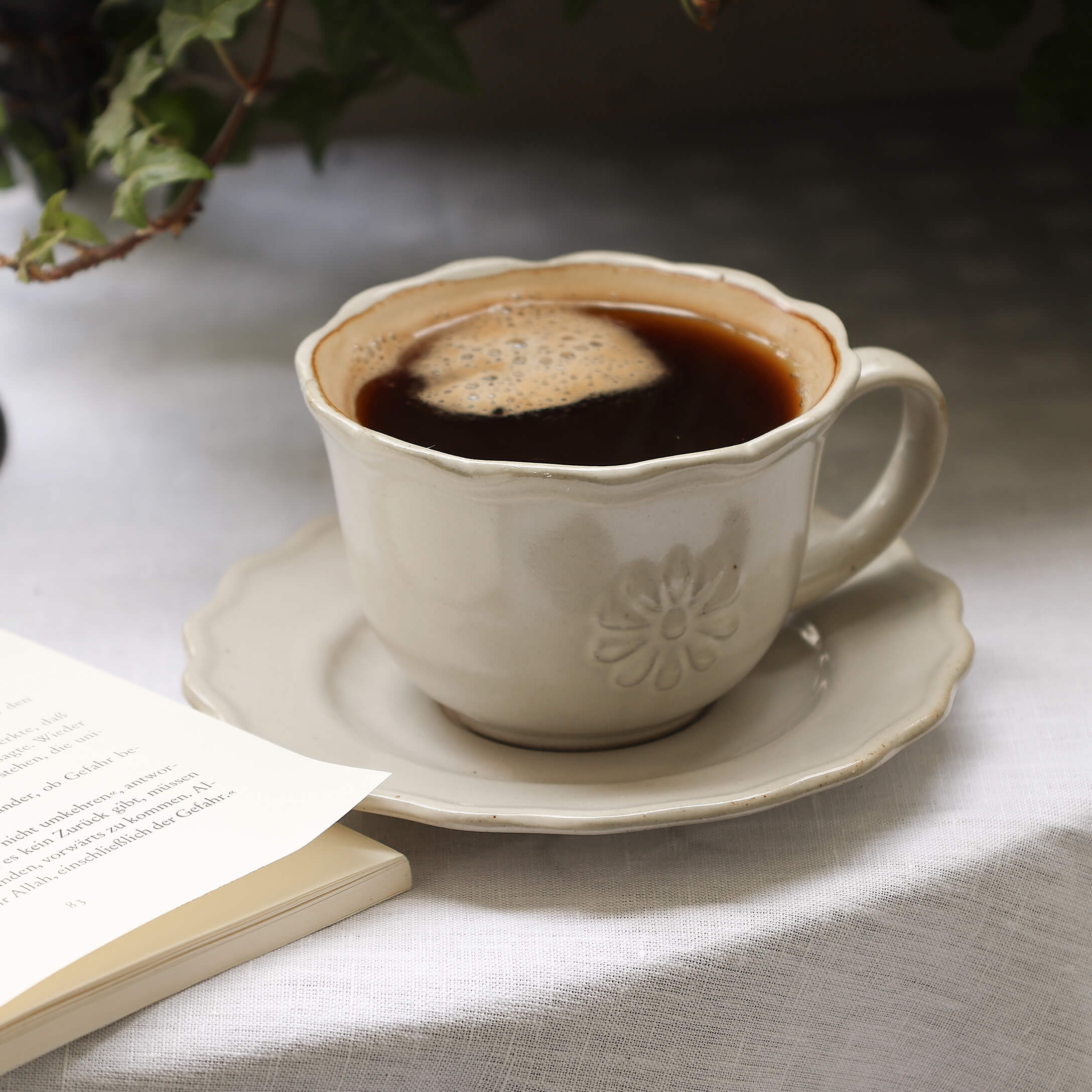 Beige Keramiktasse mit Kaffee auf Untertasse auf weissem Tischtuch neben offenem Buch