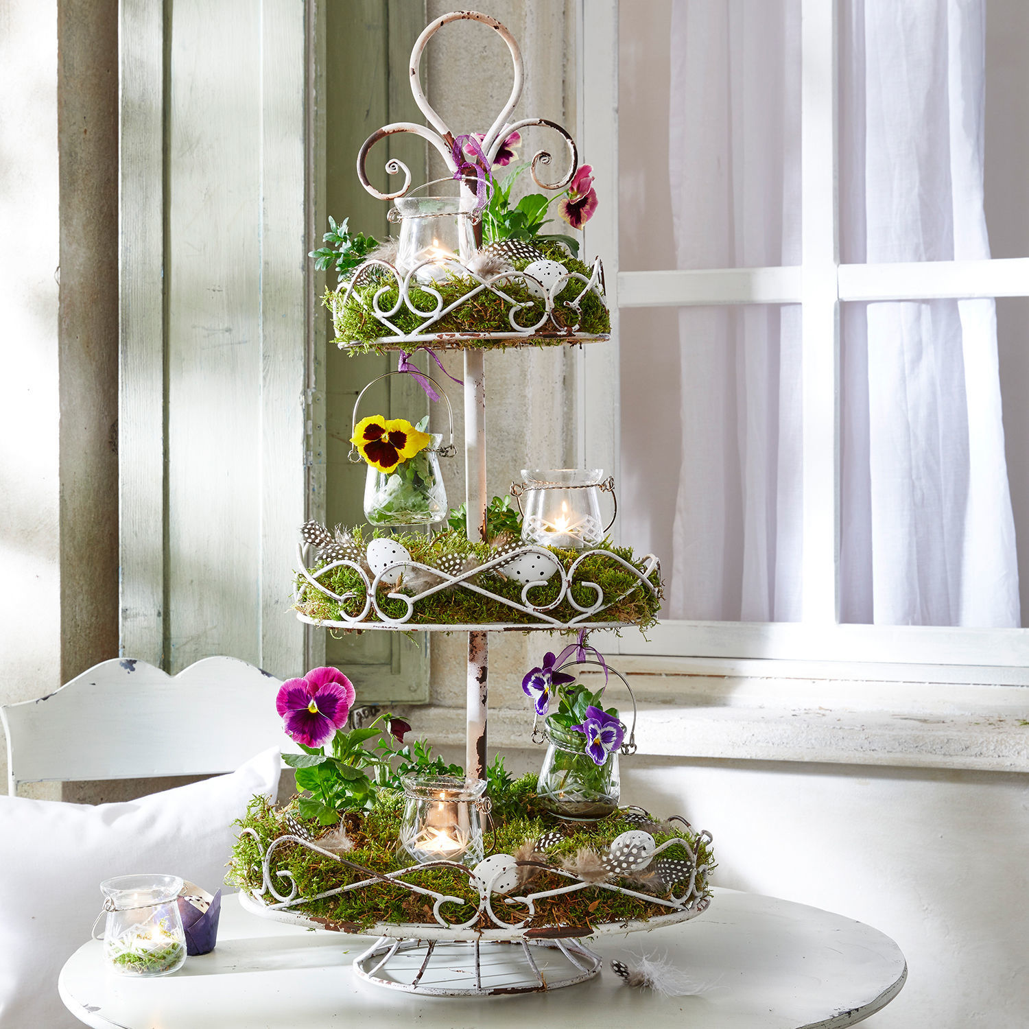 Dreistöckiges weisses Metall-Etagère mit Moos, Blumen und Kerzen als Dekoration auf weissem Tisch