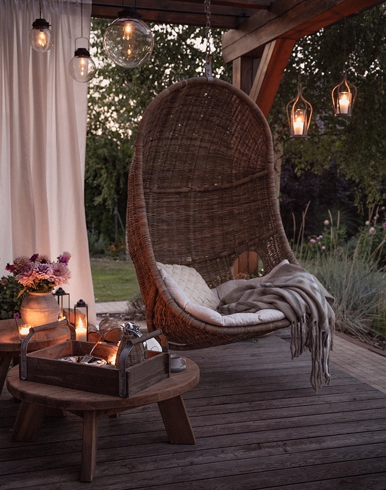 Hängesessel aus braunem Rattan auf der Terrasse mit Kissen und Decke, beleuchtet von dekorativen Lampen