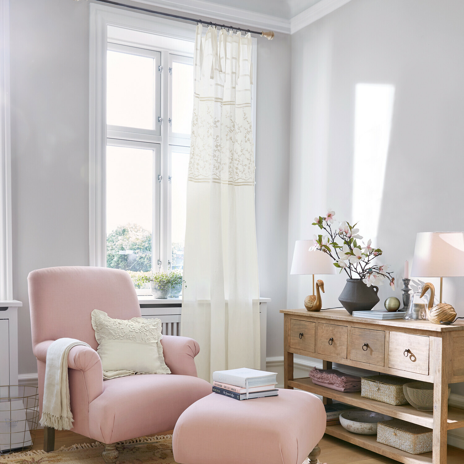 Gardine Lunérie Rosa Sessel mit Hocker und cremefarbenem Kissen in hellem Wohnzimmer mit Holzkommode und Dekorationen