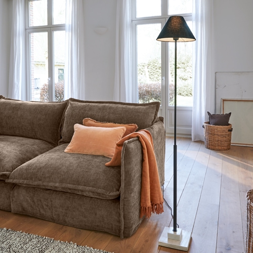 Braunes Sofa aus weichem Stoff mit orangefarbenem Kissen und Decke, schwarze Stehlampe mit Marmorfuß im hellen Wohnzimmer