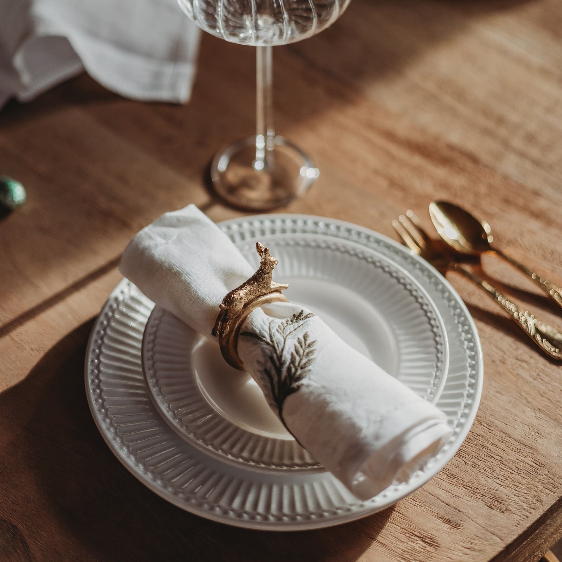 Weisser Keramikteller mit bestickter Serviette und goldenem Serviettenring auf Holztisch