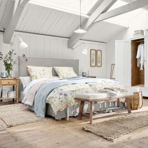Gemütliches Schlafzimmer mit hellgrauem Polsterbett, floraler Bettwäsche, Naturholz und Teppichen