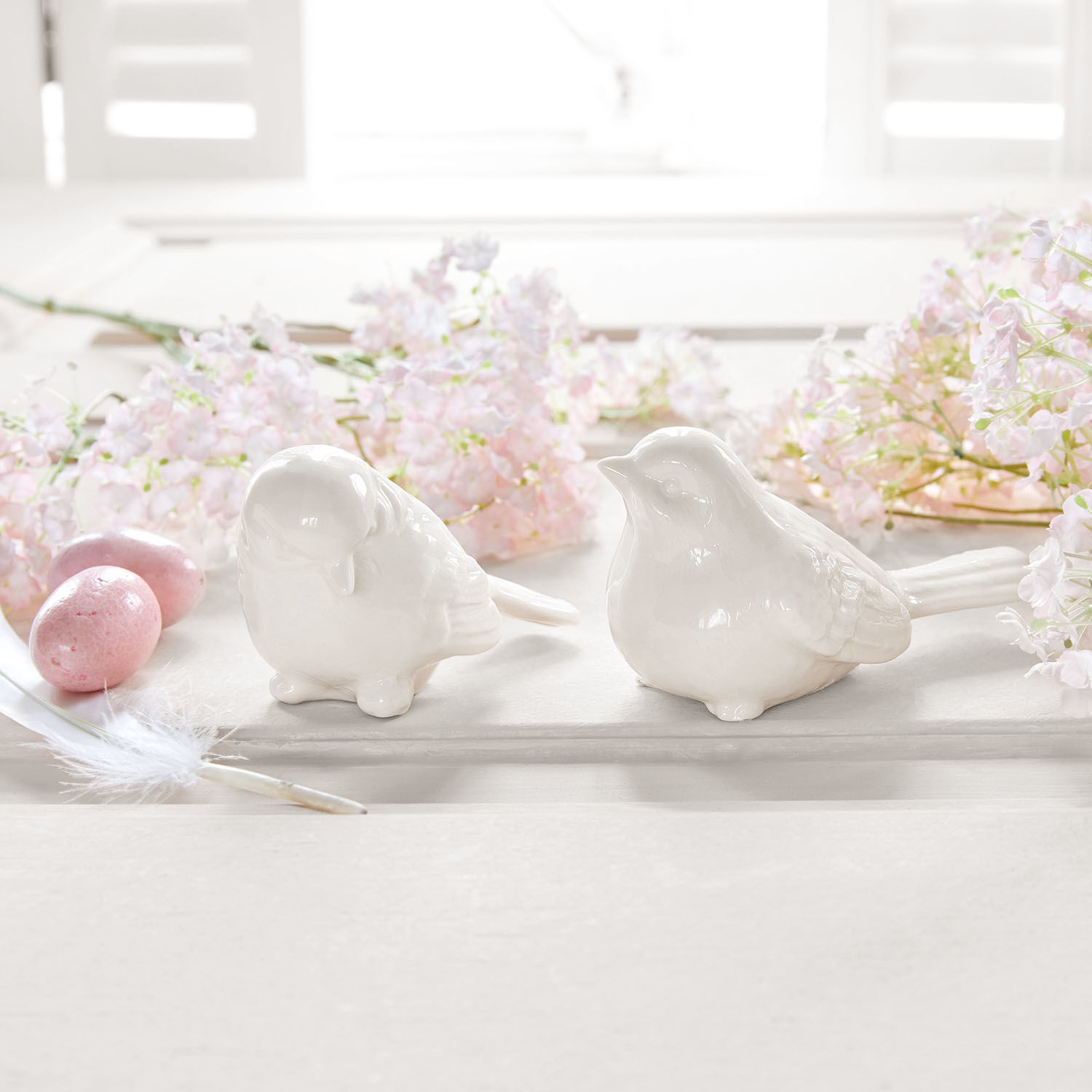 Zwei weisse Keramikvogel-Figuren auf hellem Untergrund mit rosa Blumen und Dekoeiern