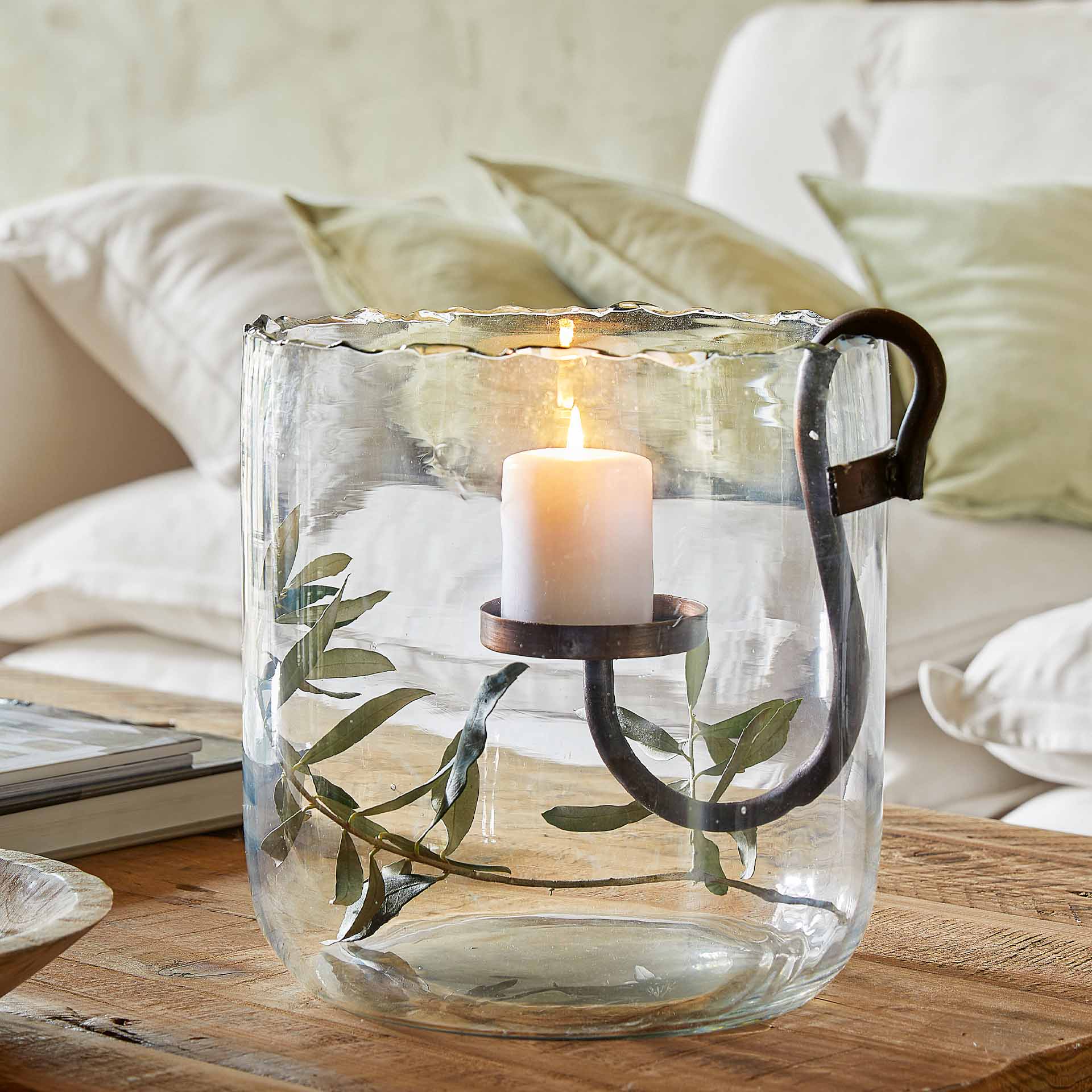 Glaswindlicht mit Metallgriff und grünen Blättern, weisse Kerze auf Holztisch