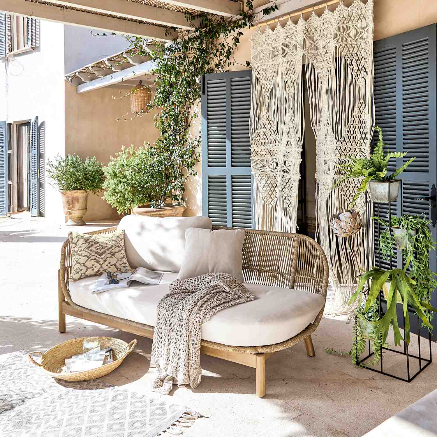 Rattansofa mit cremefarbenen Kissen und gestrickter Decke auf heller Terrasse mit Pflanzen