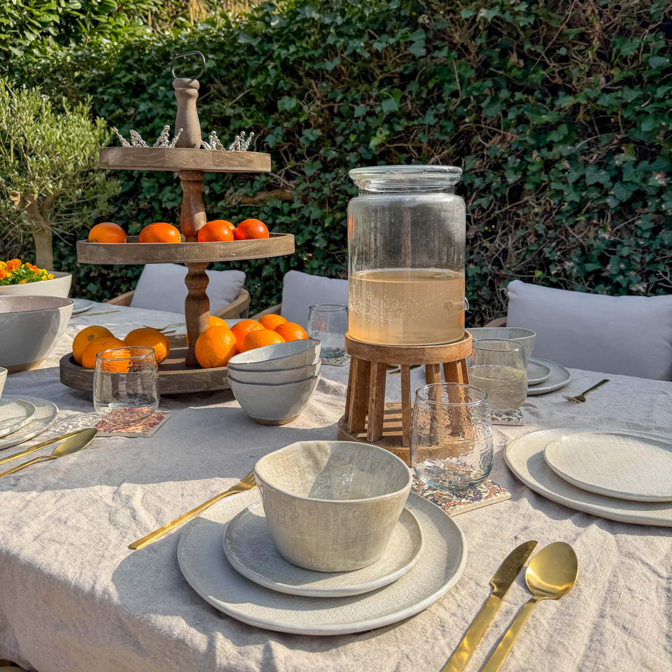 Gartentisch mit cremefarbiger Keramik, goldfarbenem Besteck und hölzernem Früchtehalter