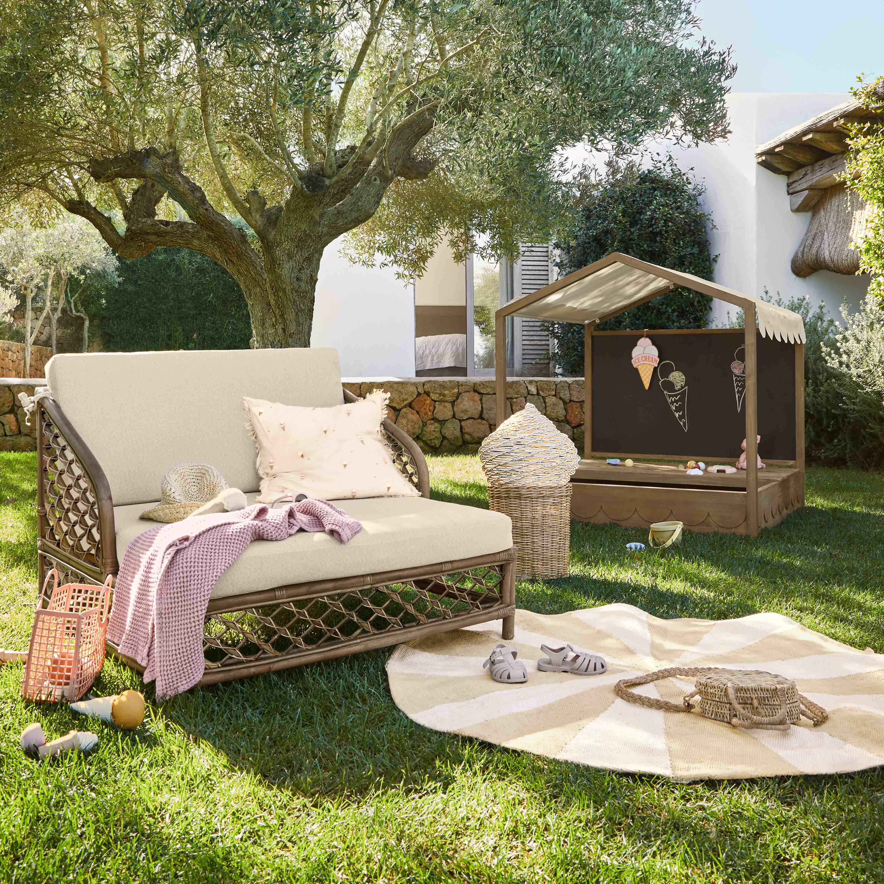 Garten mit beigem Liegestuhl, Kissen und rosa Decke auf Gras unter einem Olivenbaum