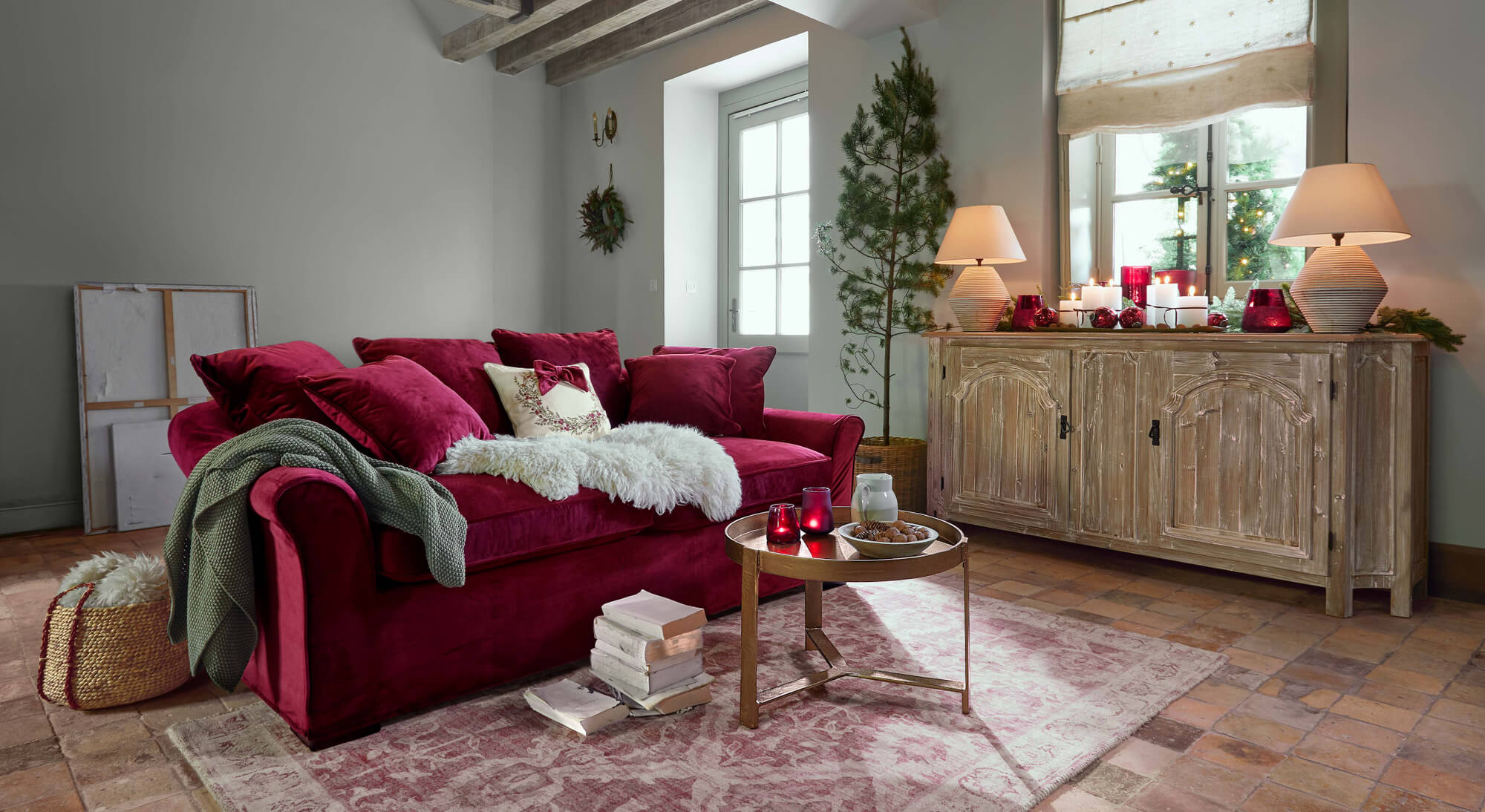 Rotes Samtsofa mit Kissen und grünem Plaid in gemütlichem Wohnzimmer mit Holzkommode und Weihnachtsdekoration