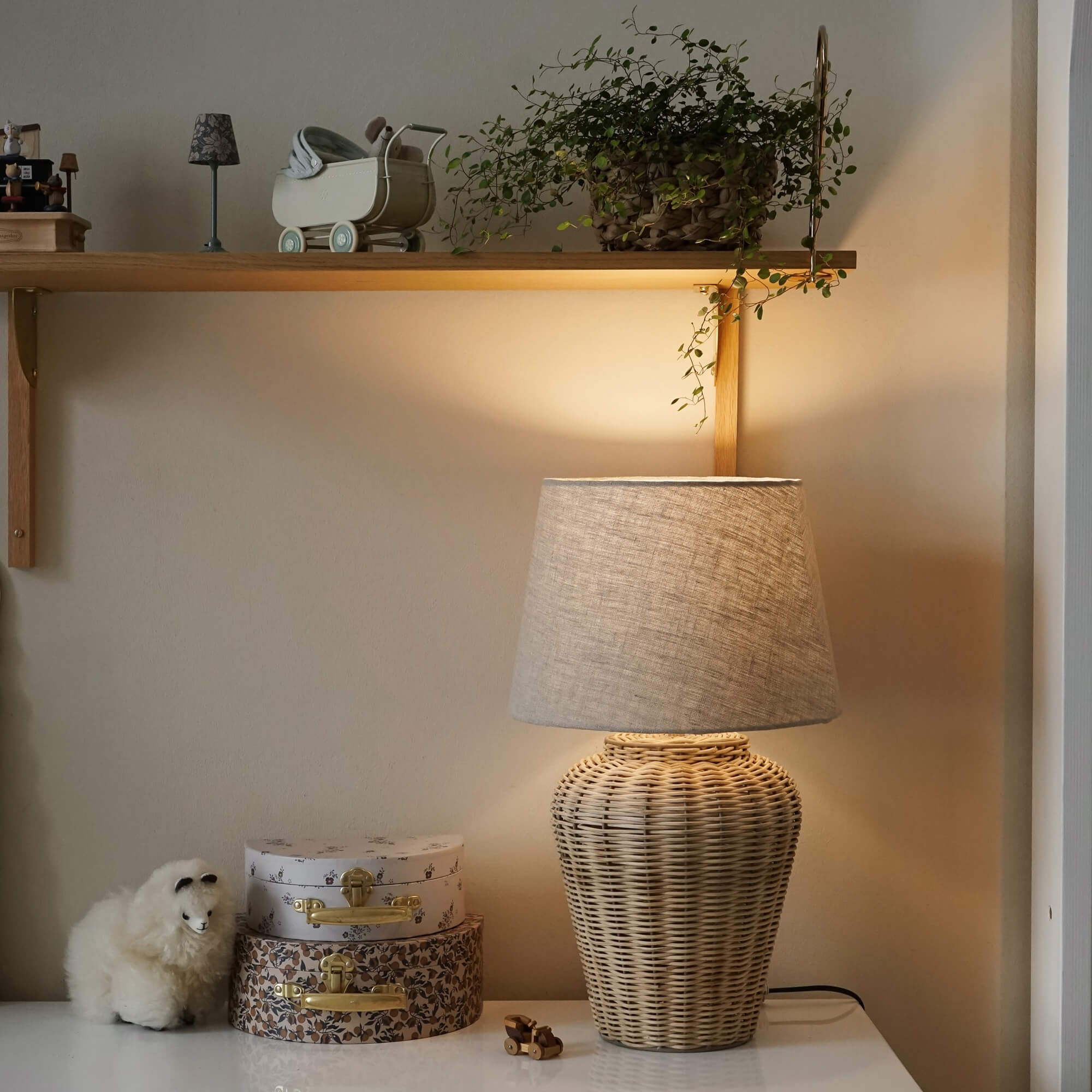 Tischlampe aus geflochtenem Rattan mit beigem Stoffschirm auf weissem Tisch mit Dekoration und Pflanze