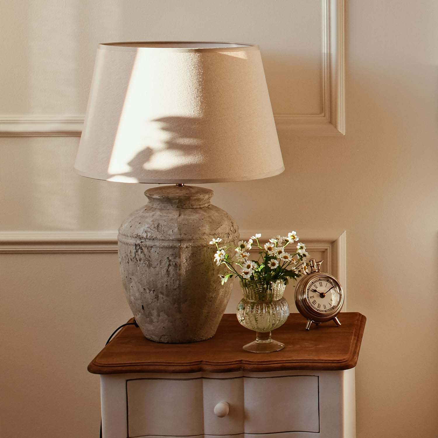 Tischlampe mit beigem Schirm und Keramikfuß im Vintage-Stil auf Holztisch mit Blumen und Wecker