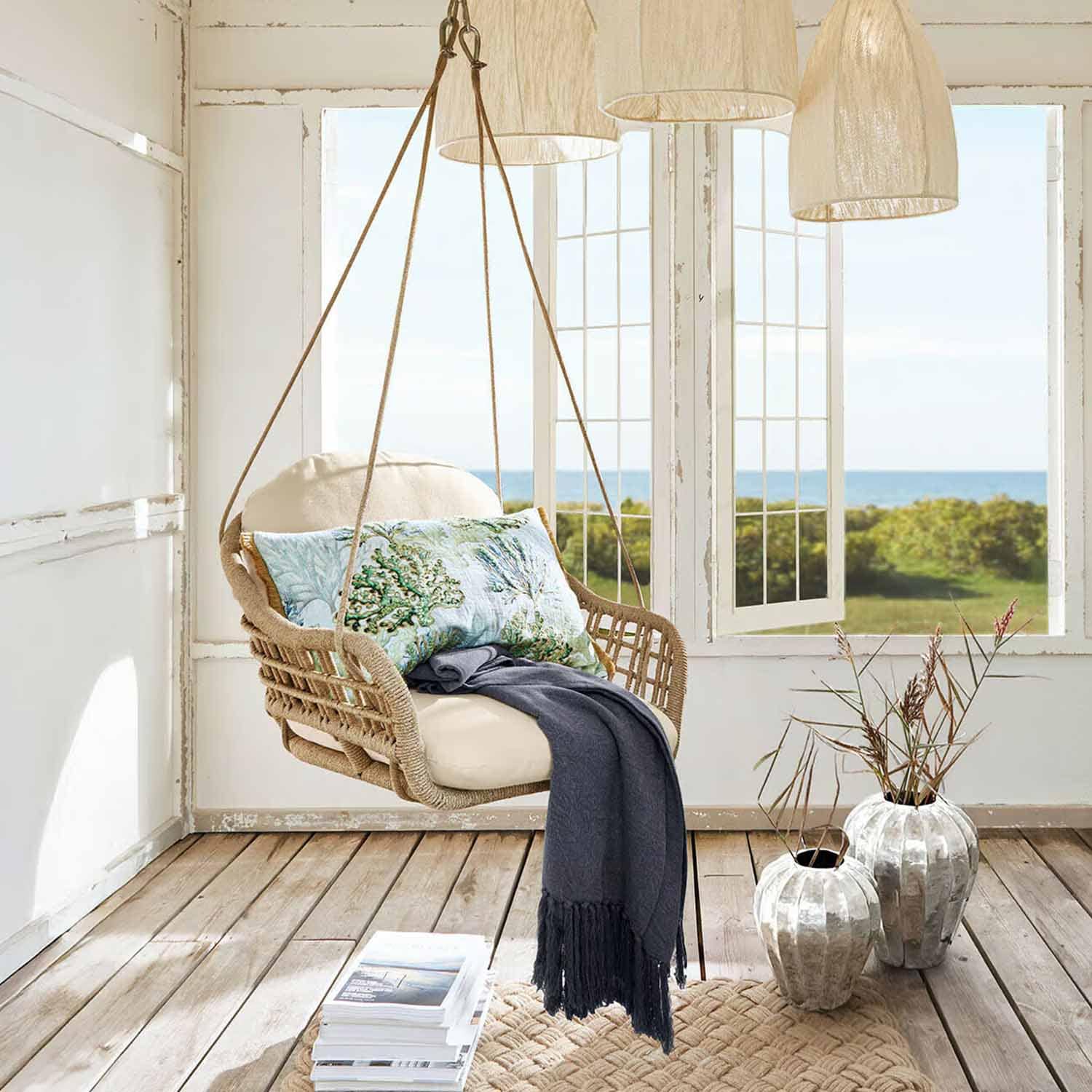 Hängesessel aus Rattan mit cremefarbenem Kissen und blauem Plaid in hellem Raum mit Meerblick