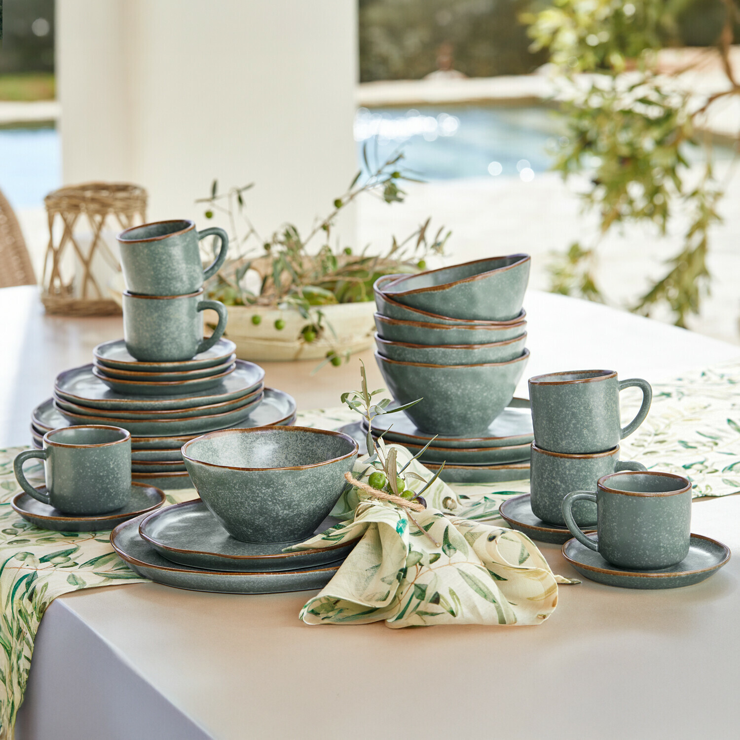 Grünes Steingut-Geschirrset mit Tassen und Tellern auf hellem Tisch mit dekorativem Stoff