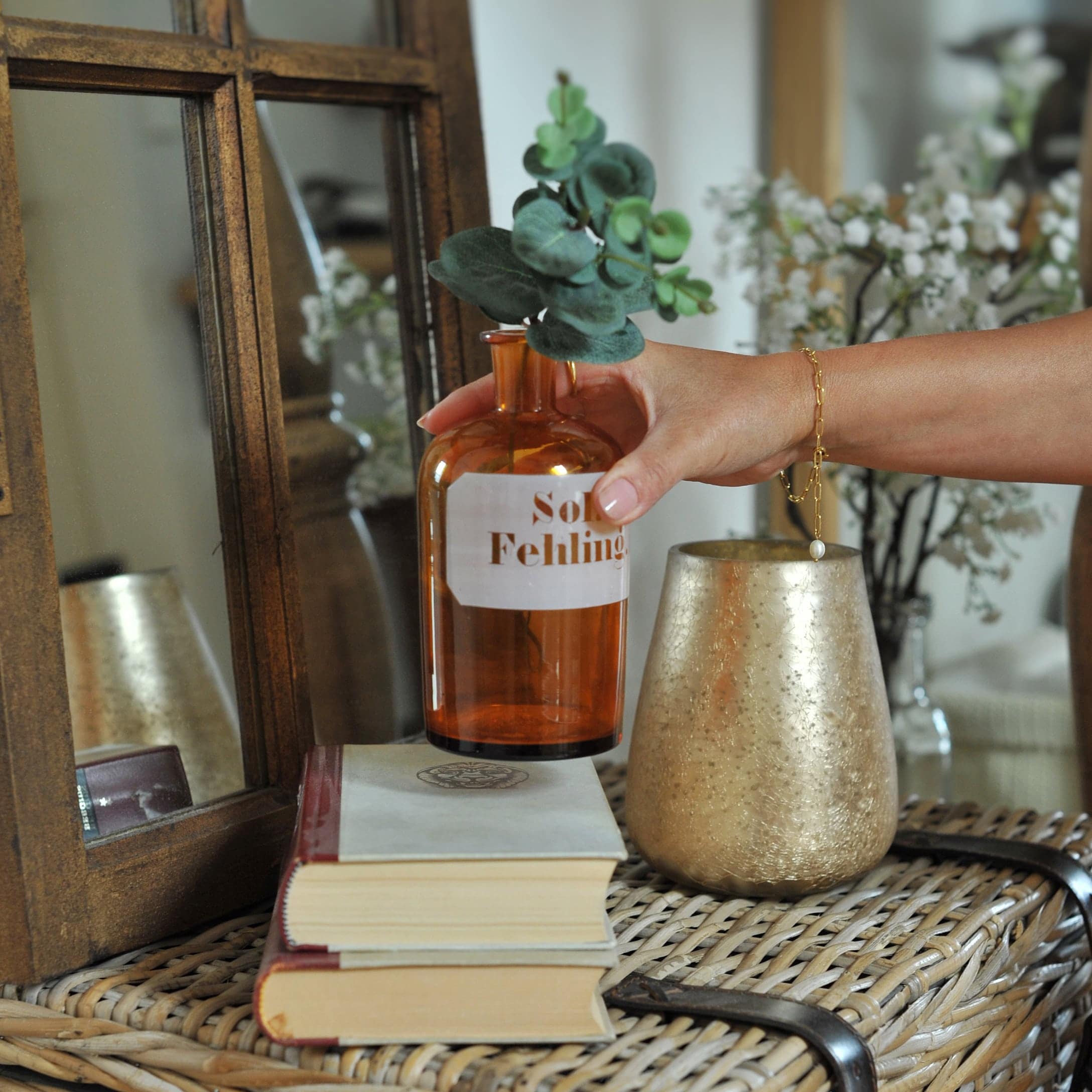 Hand hält bernsteinfarbene Glasflasche mit grünen Blättern auf Rattantisch mit Büchern und goldenem Kerzenhalter.